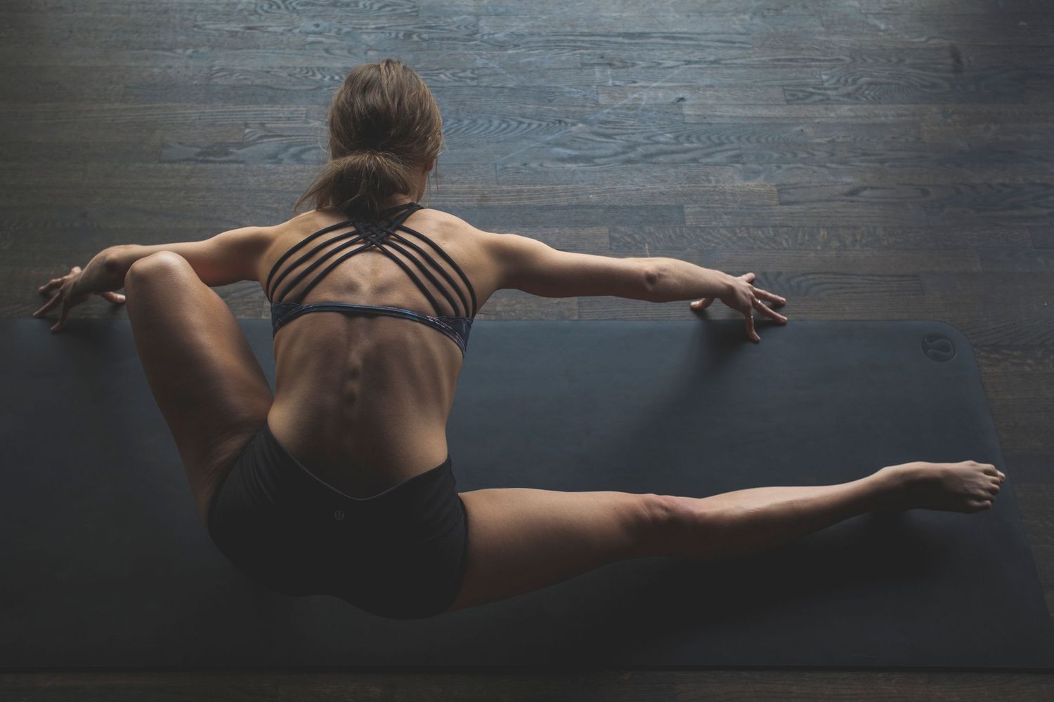 Athletic woman stretching on yoga mat in dark studio, demonstrating flexibility in black sports bra and shorts.