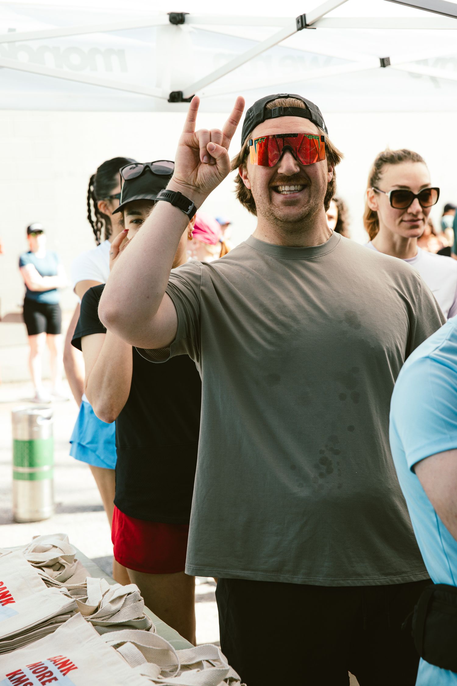 Smiling man in cap and red sunglasses making rock sign at an outdoor athletic event under a tent.