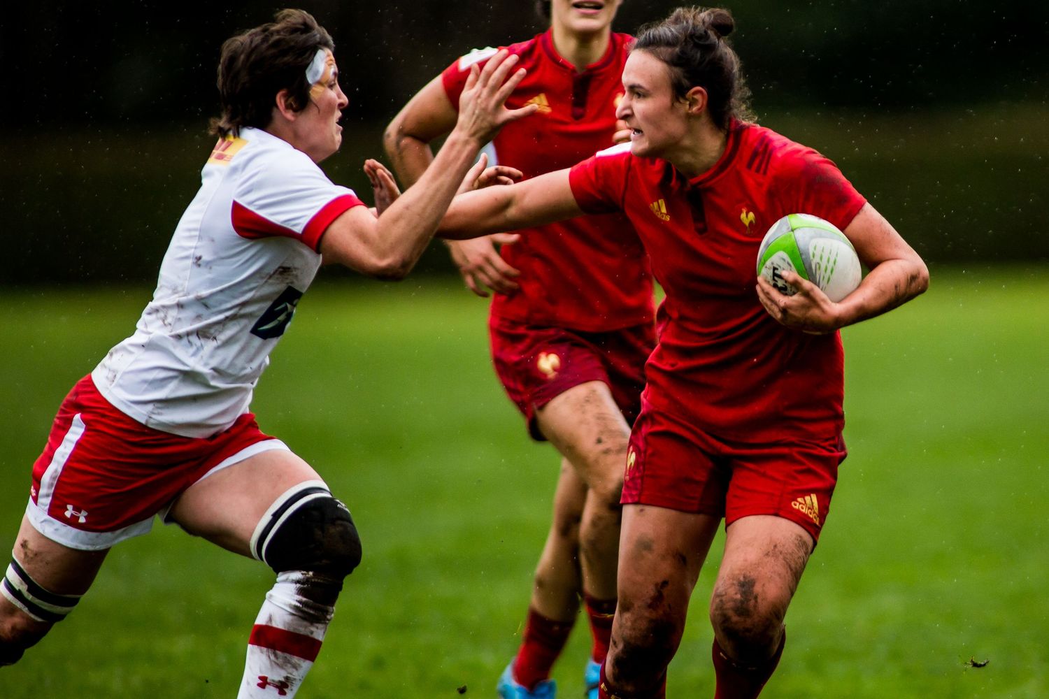 Women rugby players in red jerseys compete intensely on a green field during a match.