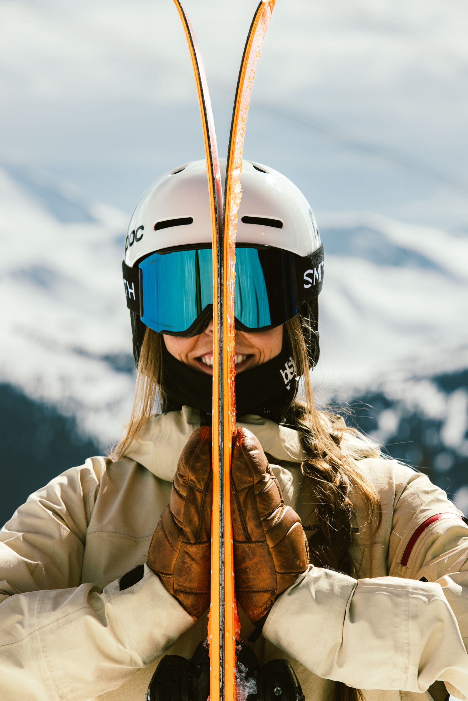 Smiling female skier holding orange Black Crows skis wearing white POC helmet and Smith goggles with snowy mountains behind.