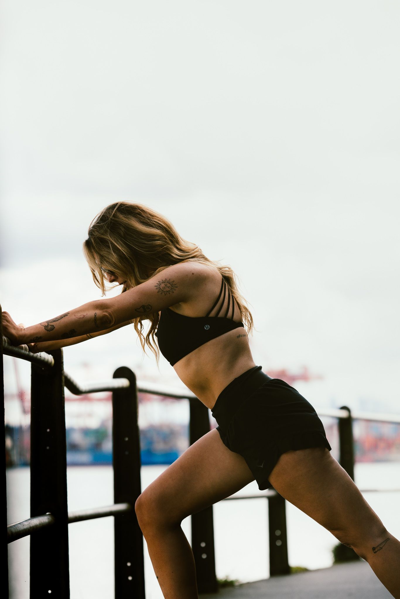 Fit woman in black sports bra and shorts leaning on railing by waterfront stretching.