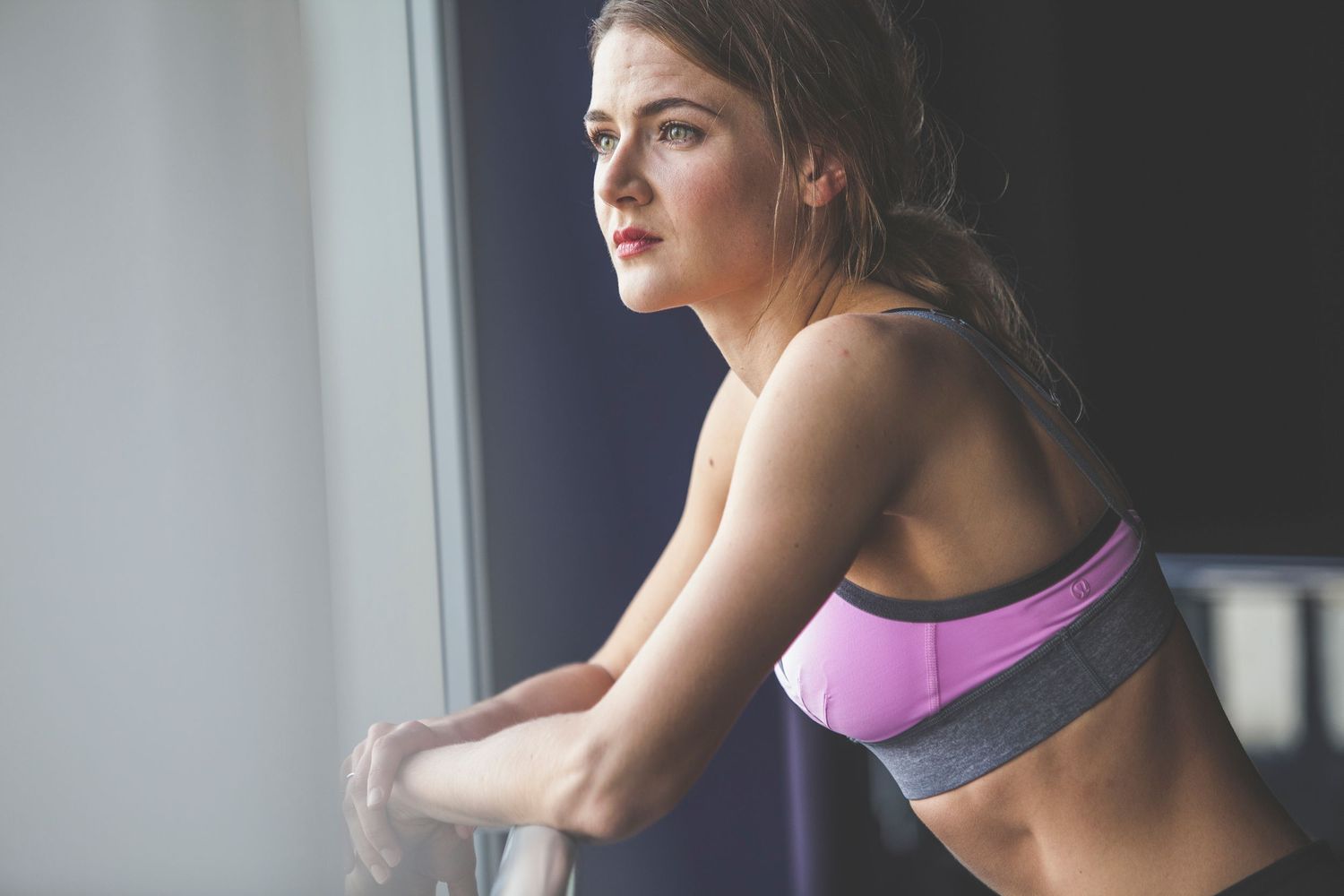 A fit young woman in a pink sports bra leans against a window, gazing outside after a workout.