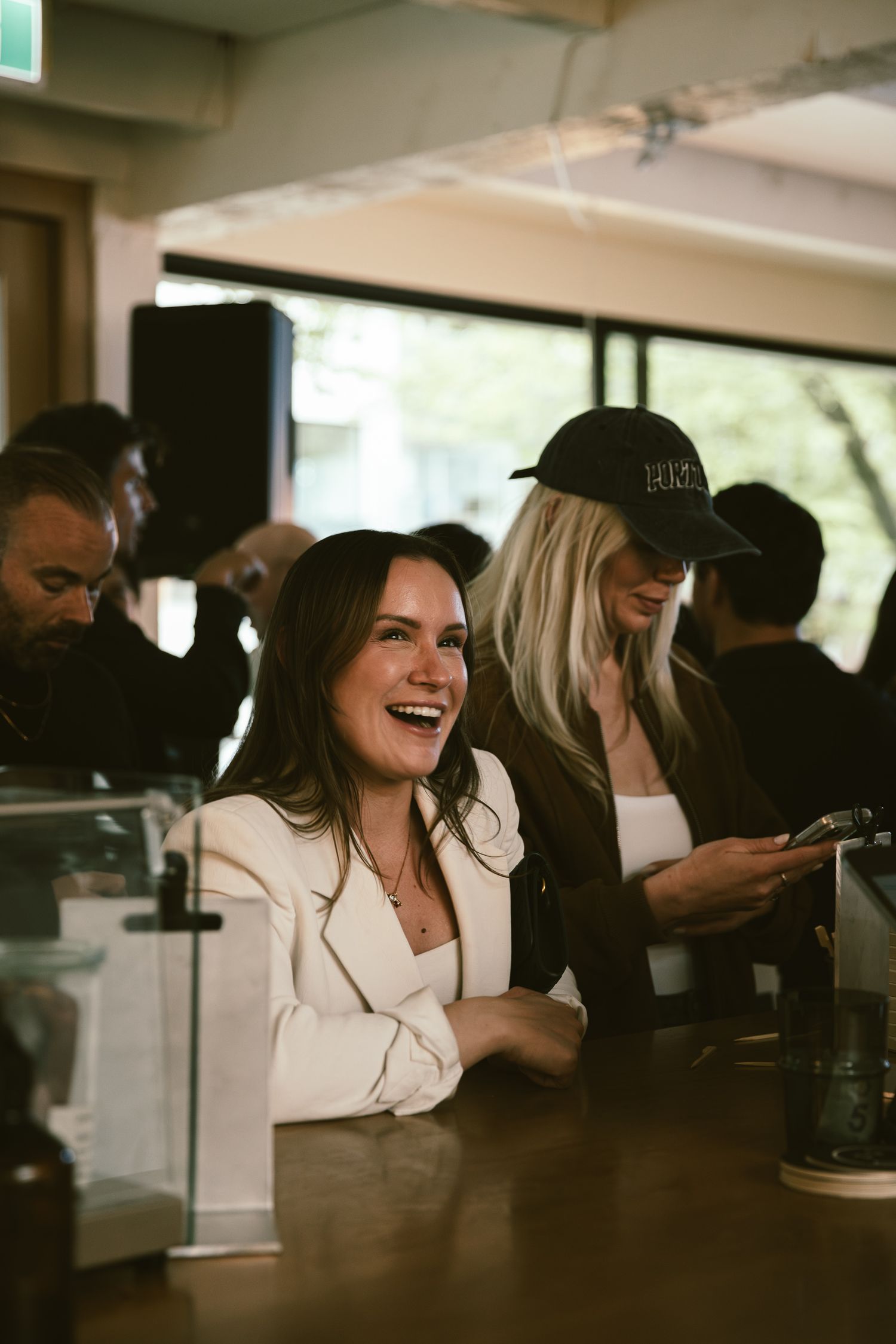 A laughing woman in a white blazer sits at a bar counter with friends in a busy cafe.