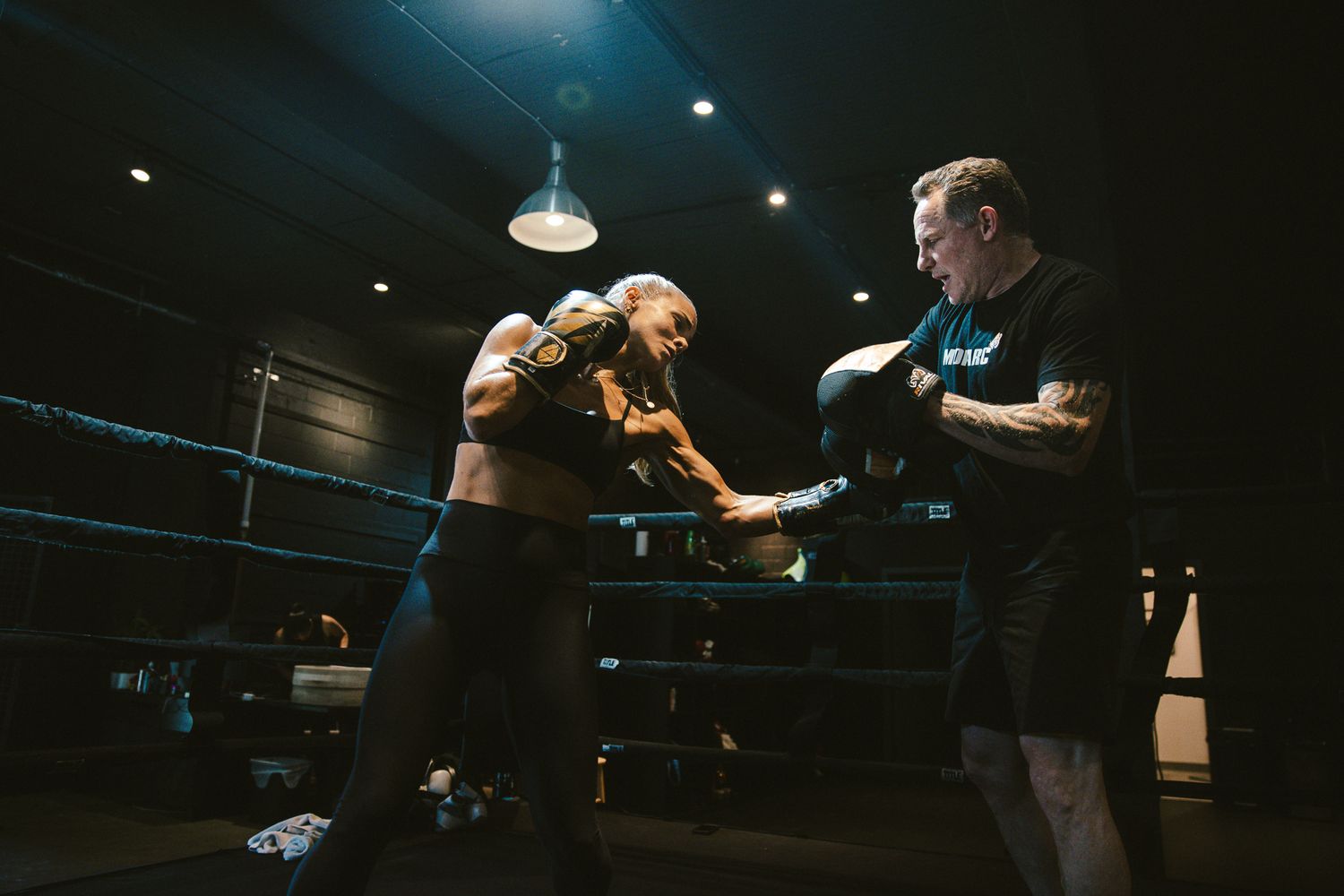 Two men sparring in a dark boxing ring during an intense training session.