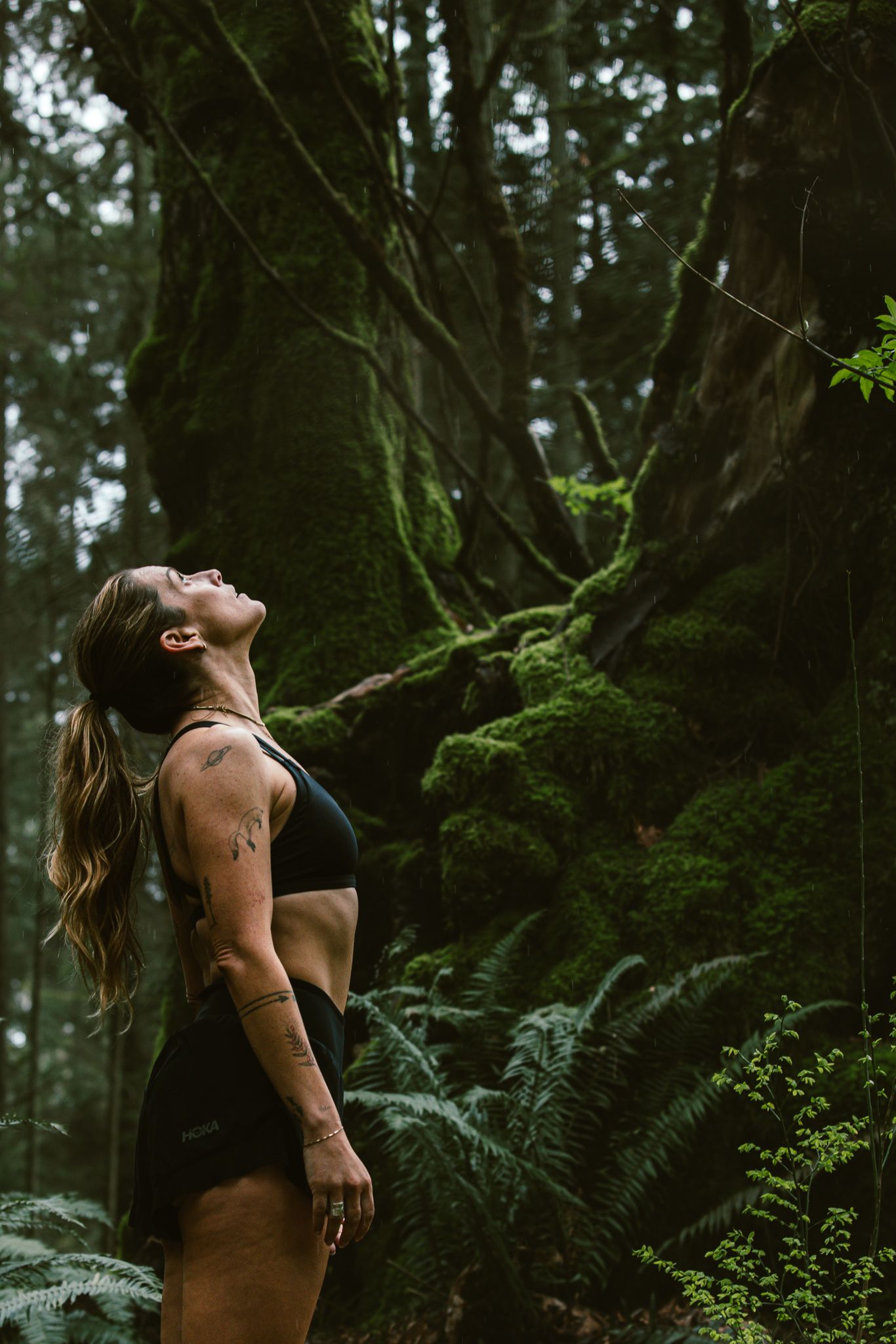 Athletic woman in black sports bra and shorts standing in lush green forest looking up.