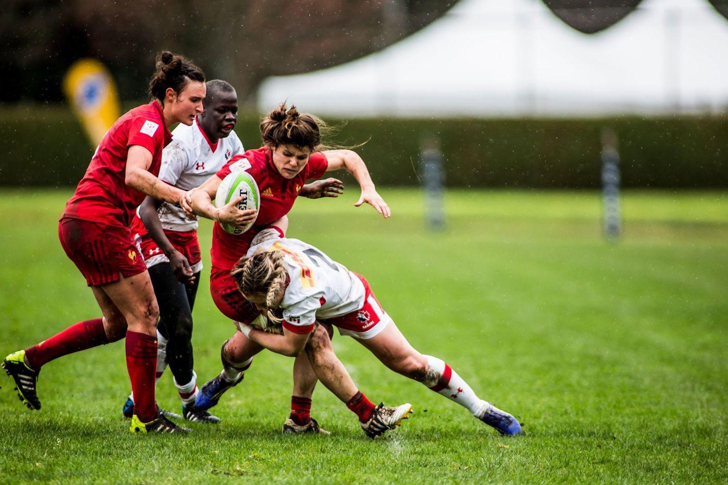 Women rugby players in red jerseys compete in a tackle during an outdoor match on a green field.