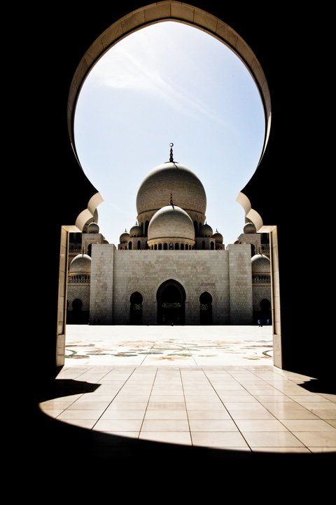 View through ornate arched gateway of Sheikh Zayed Grand Mosque white domes and minarets under blue sky in Abu Dhabi.