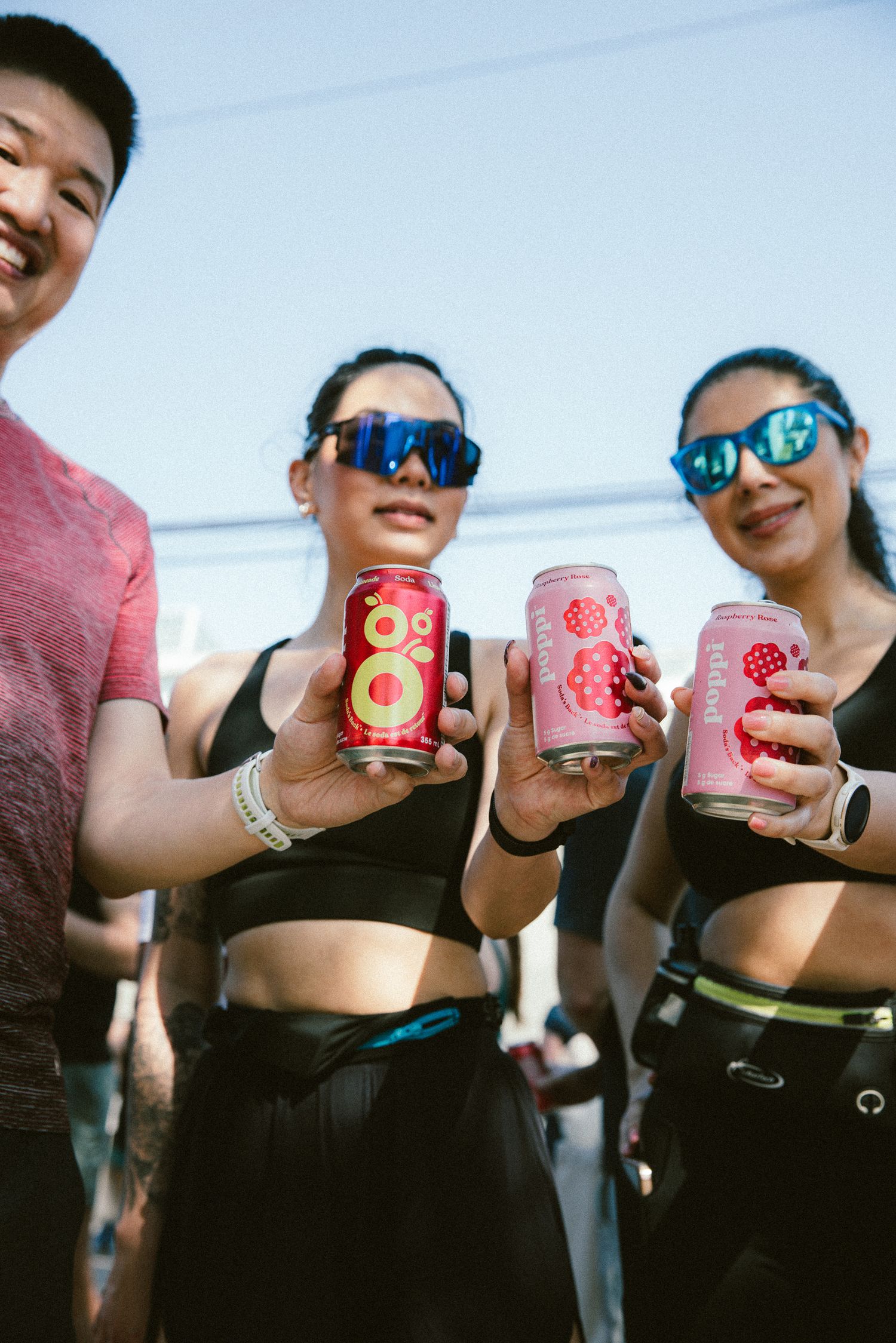 Three smiling friends in athletic wear holding colorful canned beverages outdoors after a run.