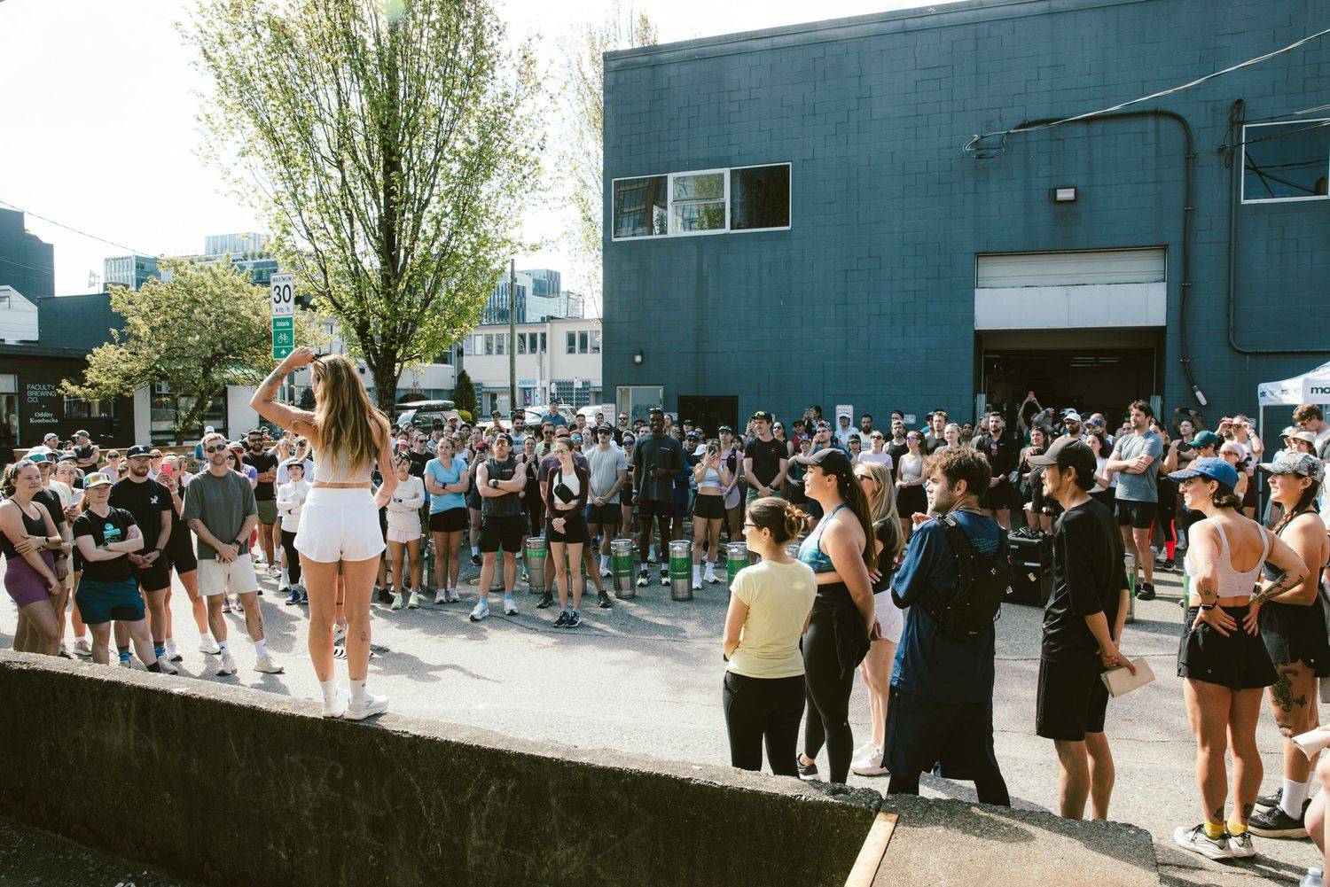 Large outdoor crowd gathered in urban area on sunny day, people standing near building watching speaker on elevated platform.
