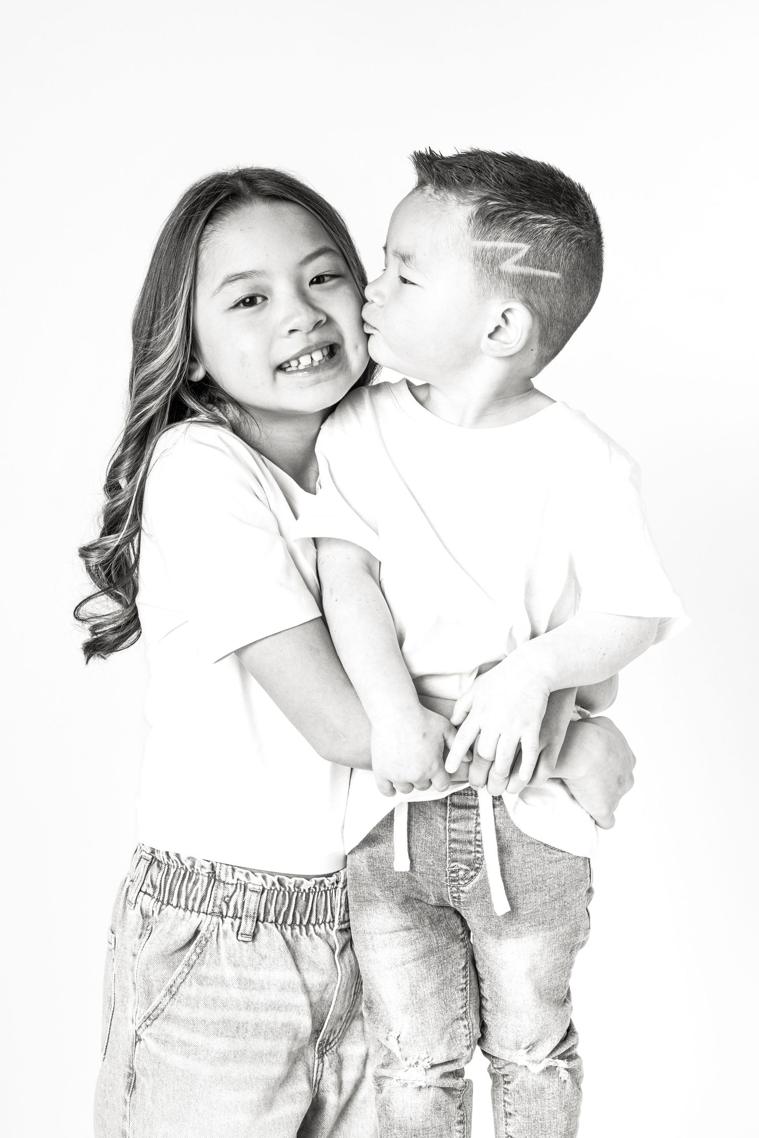 Black and white photo of a young boy kissing his smiling older sister on the cheek in studio setting.