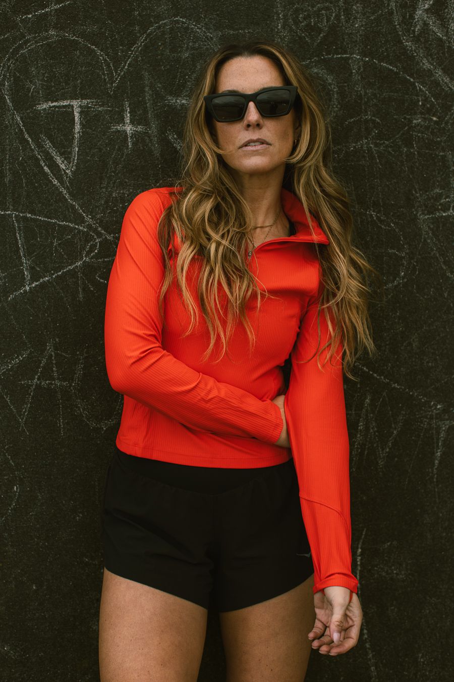 Athletic woman in a red zip-up top and black shorts posing against a dark graffiti wall with sunglasses.