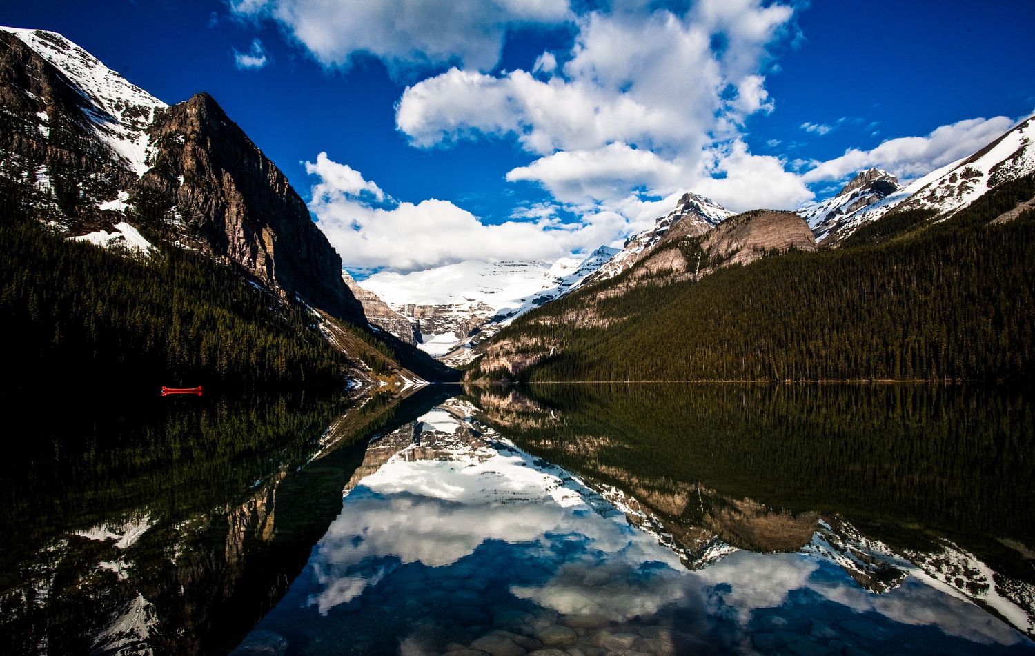 Serene Lake Louise reflects snow-capped Rocky Mountains and evergreen forests under a blue sky with clouds in Banff, Canada.
