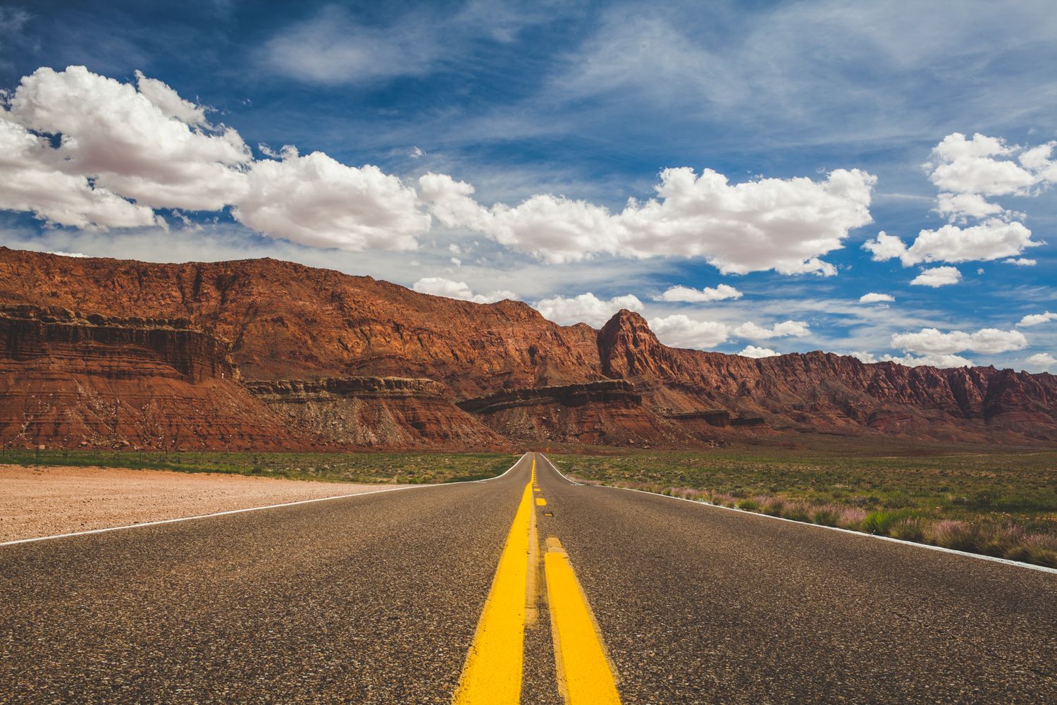A long straight desert highway with yellow center lines leads toward dramatic red rock cliffs under a blue sky with white clouds.