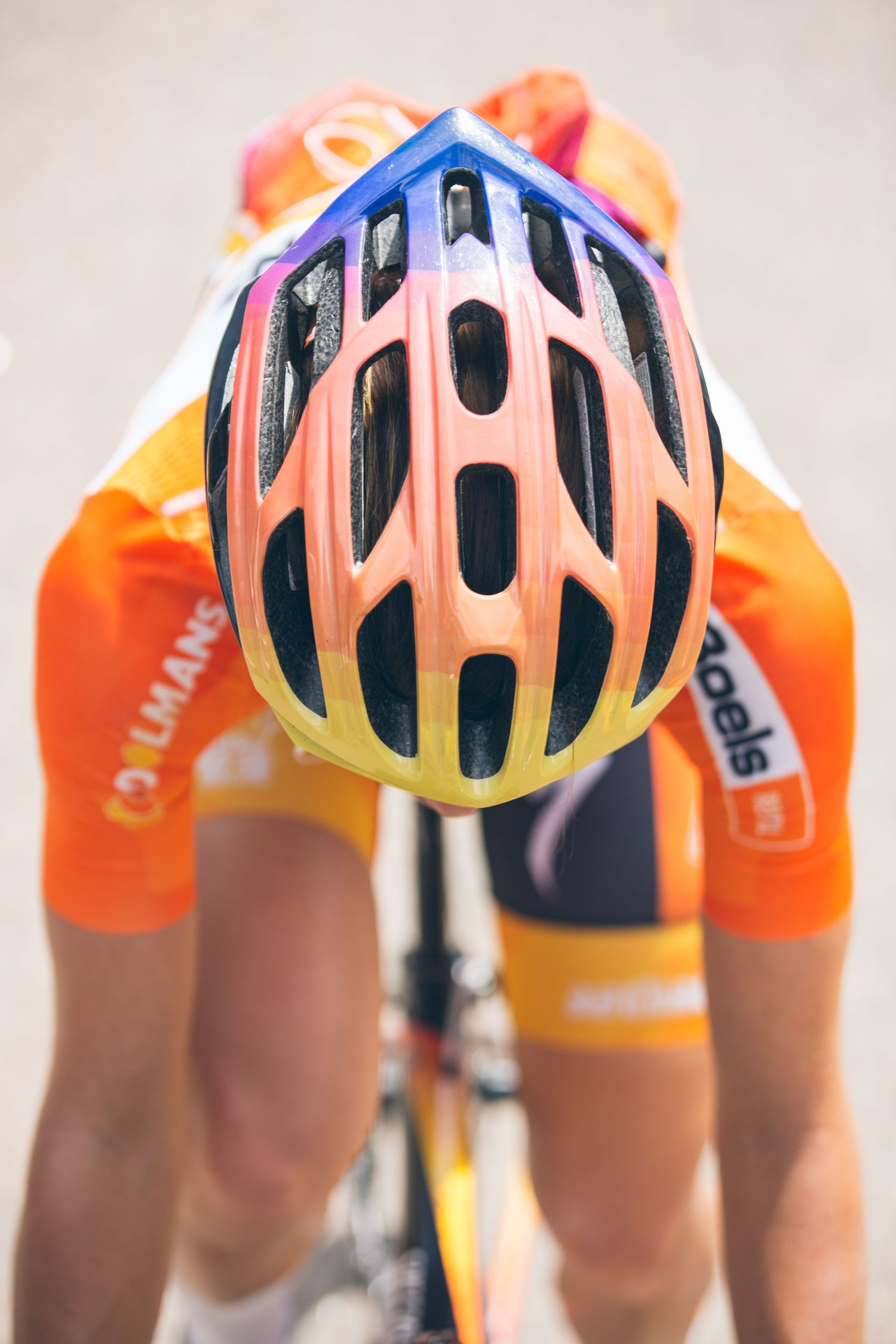 A cyclist in an orange team jersey wears a colorful rainbow-gradient helmet, leaning forward on their bike during a race.