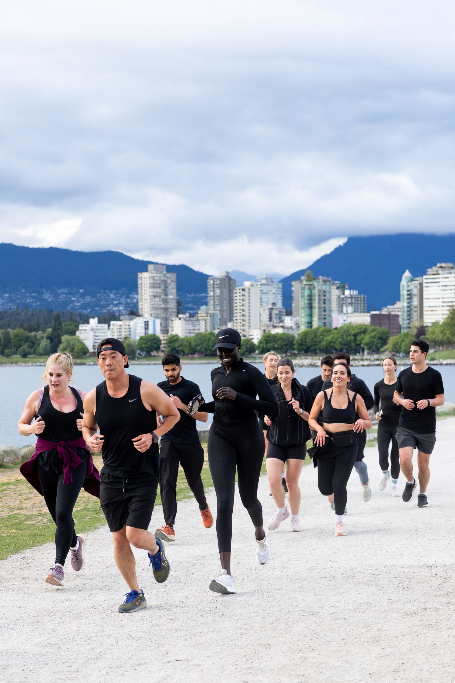 Group of runners in black athletic wear jogging along a waterfront path with city skyline and mountains behind.