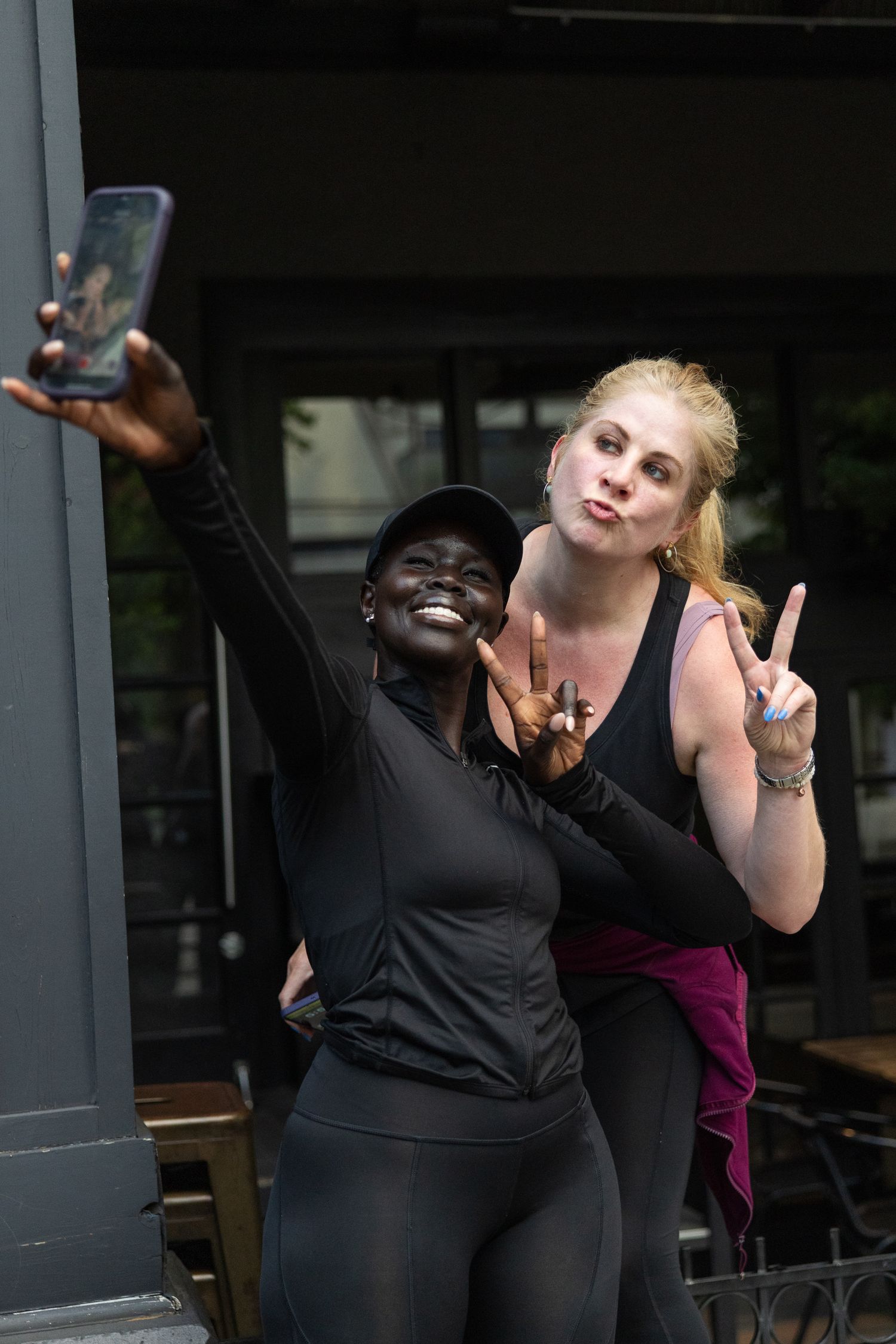 Two women in black athletic wear take a selfie outdoors, flashing peace signs and smiling.