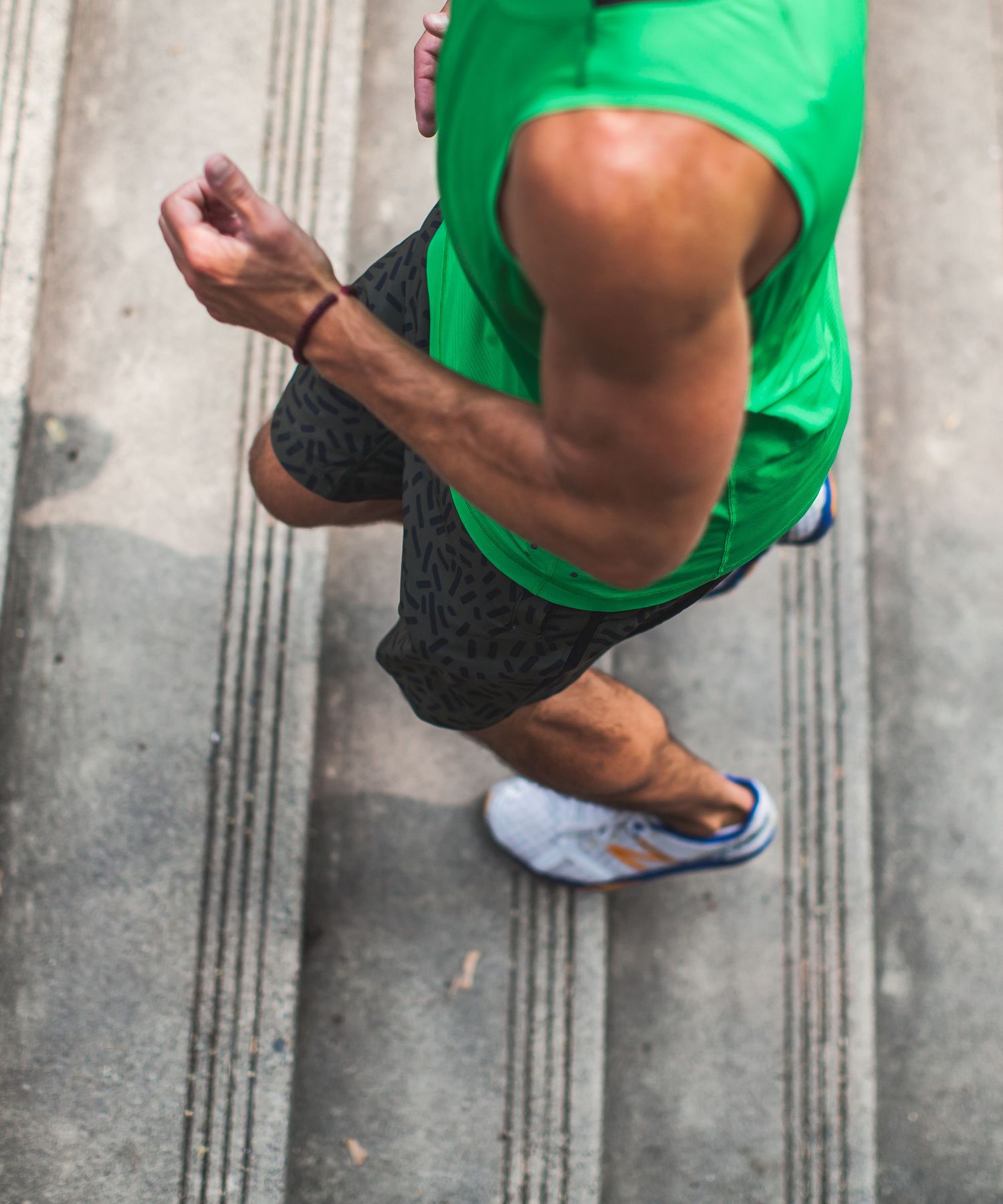 Aerial view of a muscular man in a green tank top and black shorts running on a concrete surface wearing blue sneakers.
