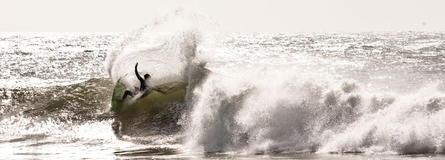 A surfer rides a powerful crashing wave in the ocean, performing an aerial maneuver with white water spray.