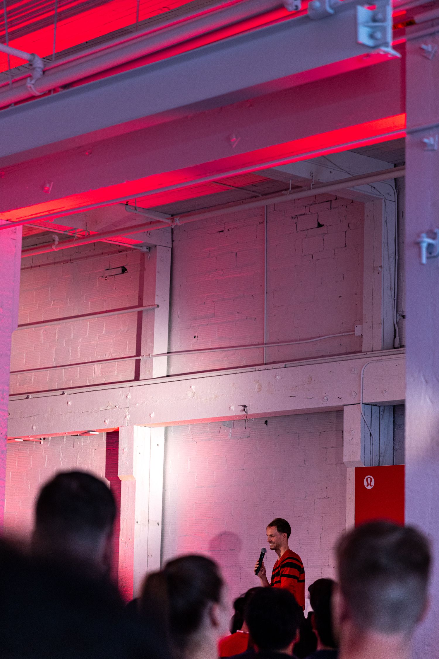 Man speaking into microphone at a Lululemon event with red neon lighting in an industrial venue.