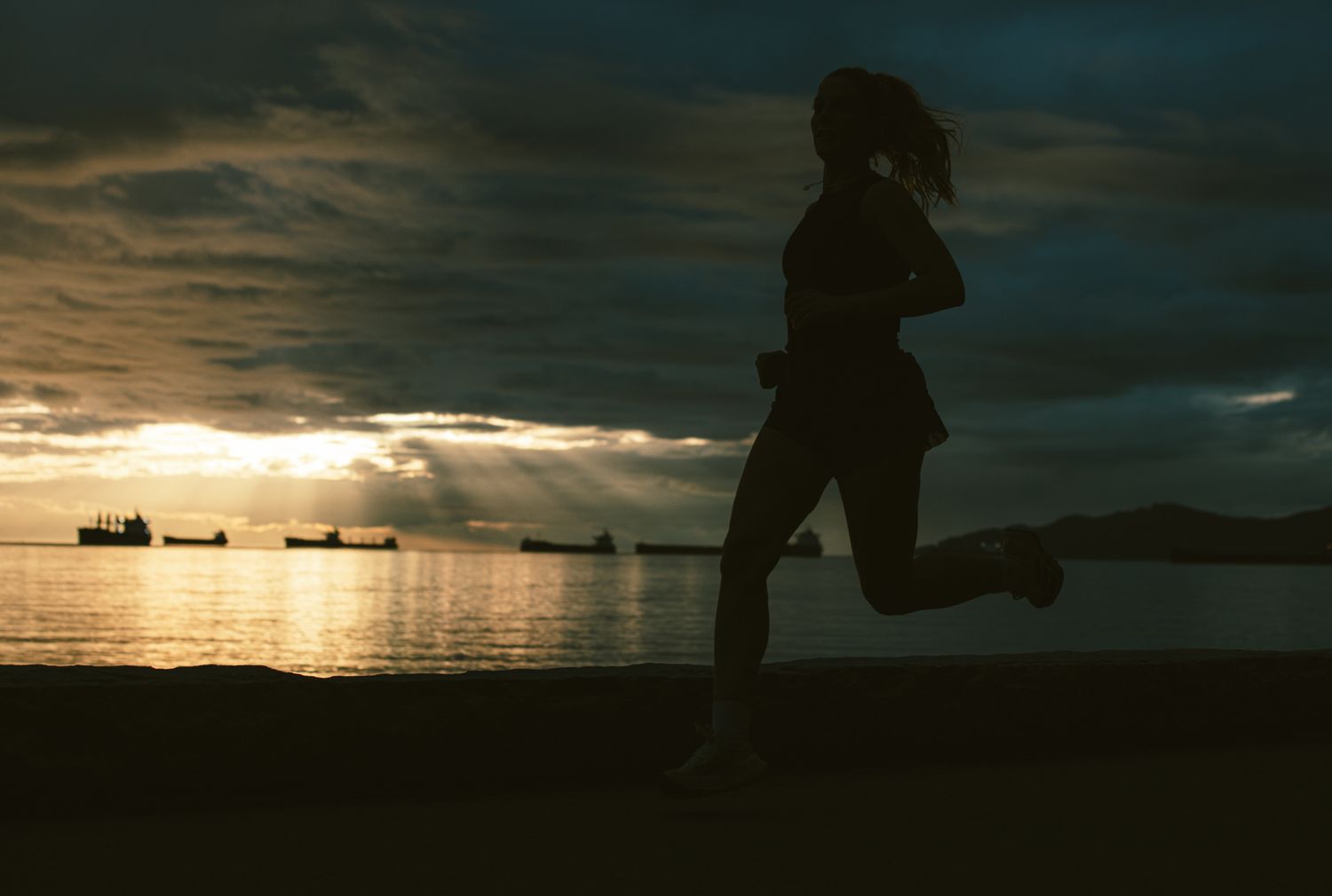 Silhouette of a female runner with a ponytail jogging by the ocean at golden sunset with ships behind.