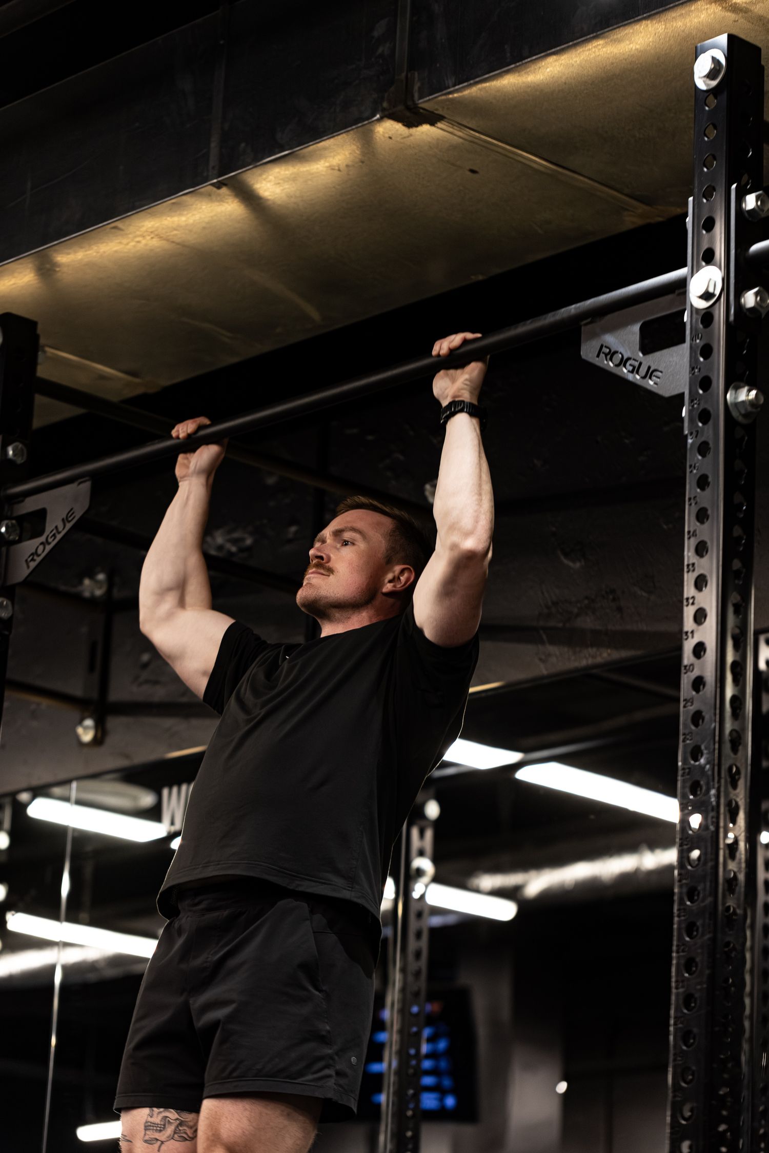 Muscular man in black performing pull-ups on a Rogue bar in a dark gym.