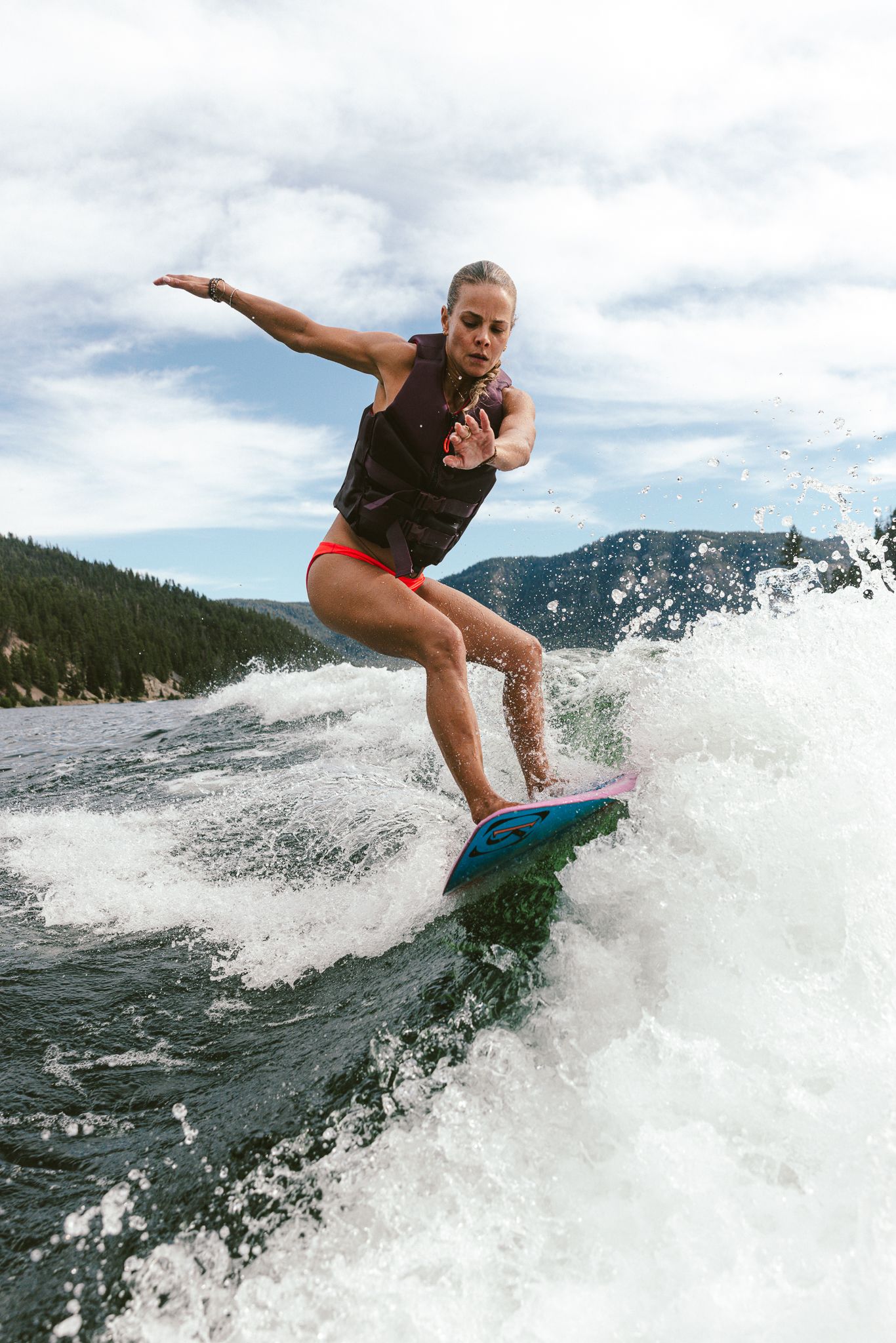 Woman wake surfing on a lake with mountain backdrop, wearing black top and red bikini bottoms.