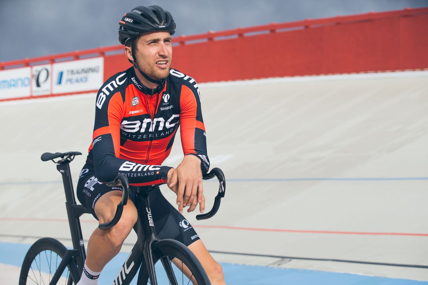Cyclist in BMC racing team jersey rests on a track bike inside a velodrome, looking confidently to the side.