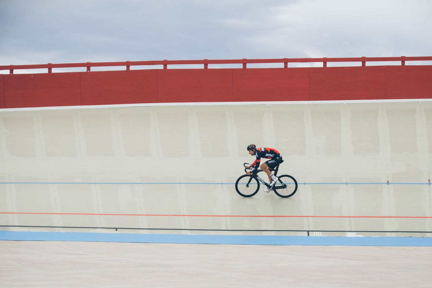 Cyclist racing on a velodrome track with red and white walls under a cloudy sky.