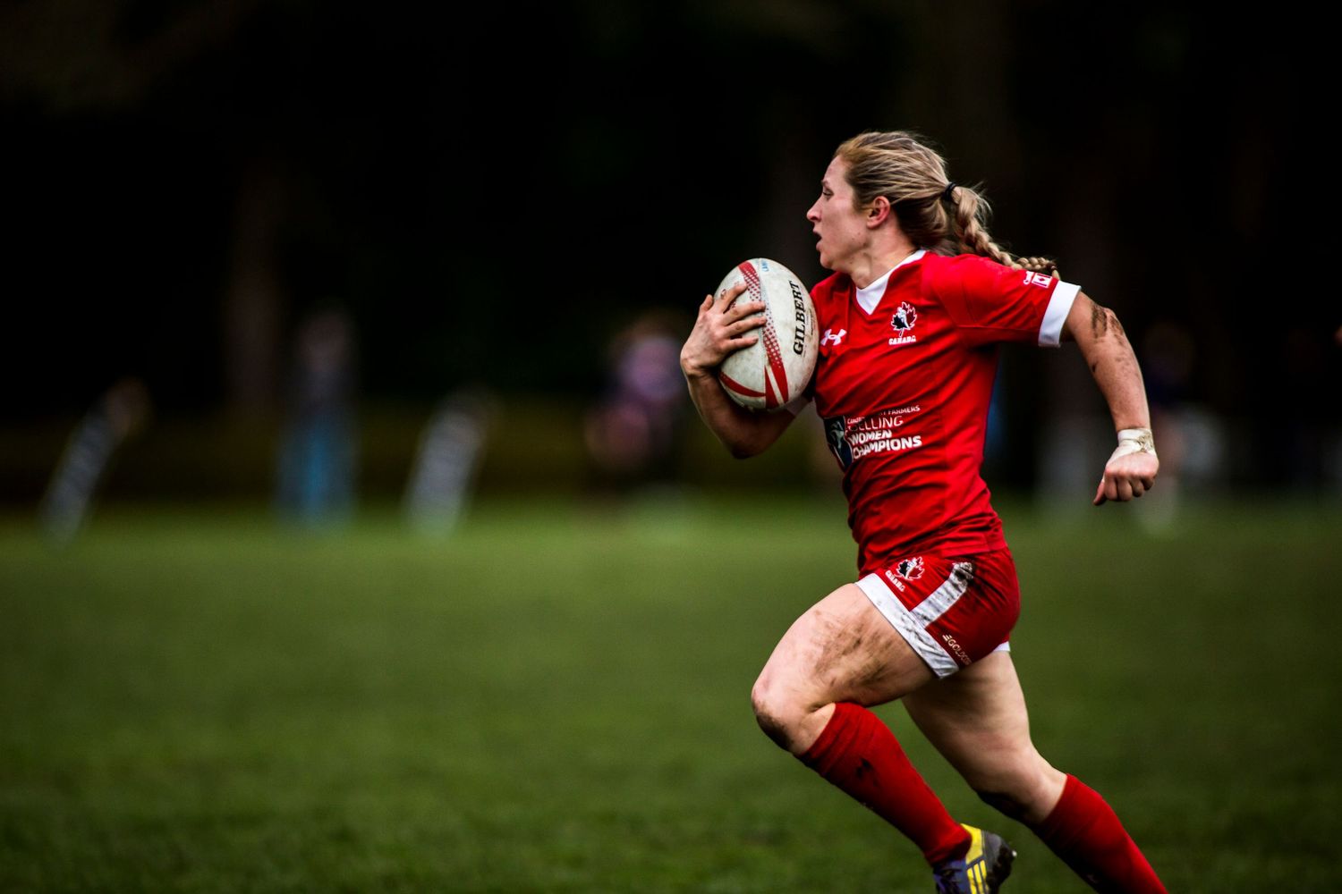 Female rugby player in red uniform sprinting across green field while carrying rugby ball during competitive match.