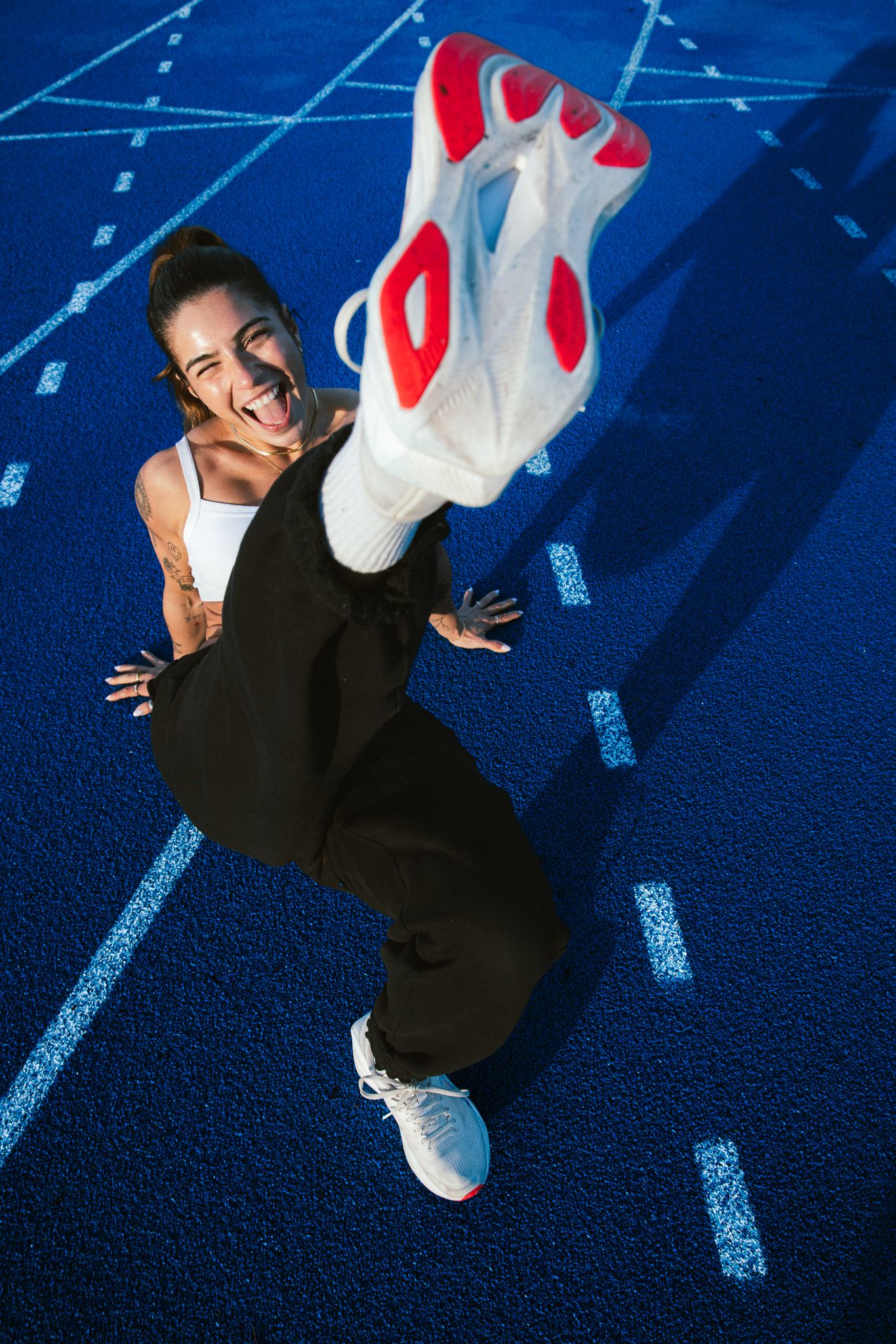 Overhead view of smiling woman kicking leg up on blue athletic track wearing white sneakers.