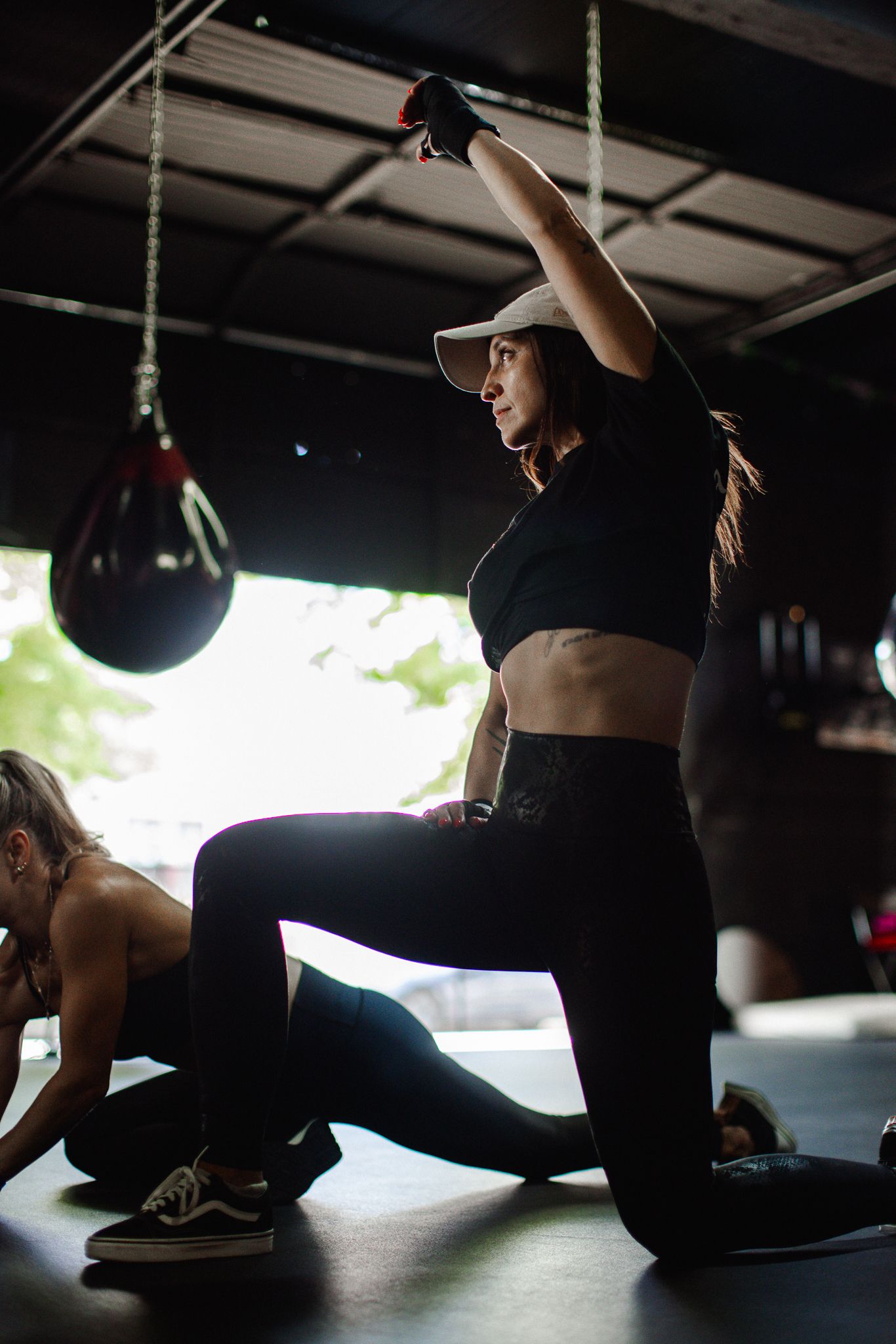 Woman in black workout gear performs a lunge with arm raised in a dark boxing gym setting.