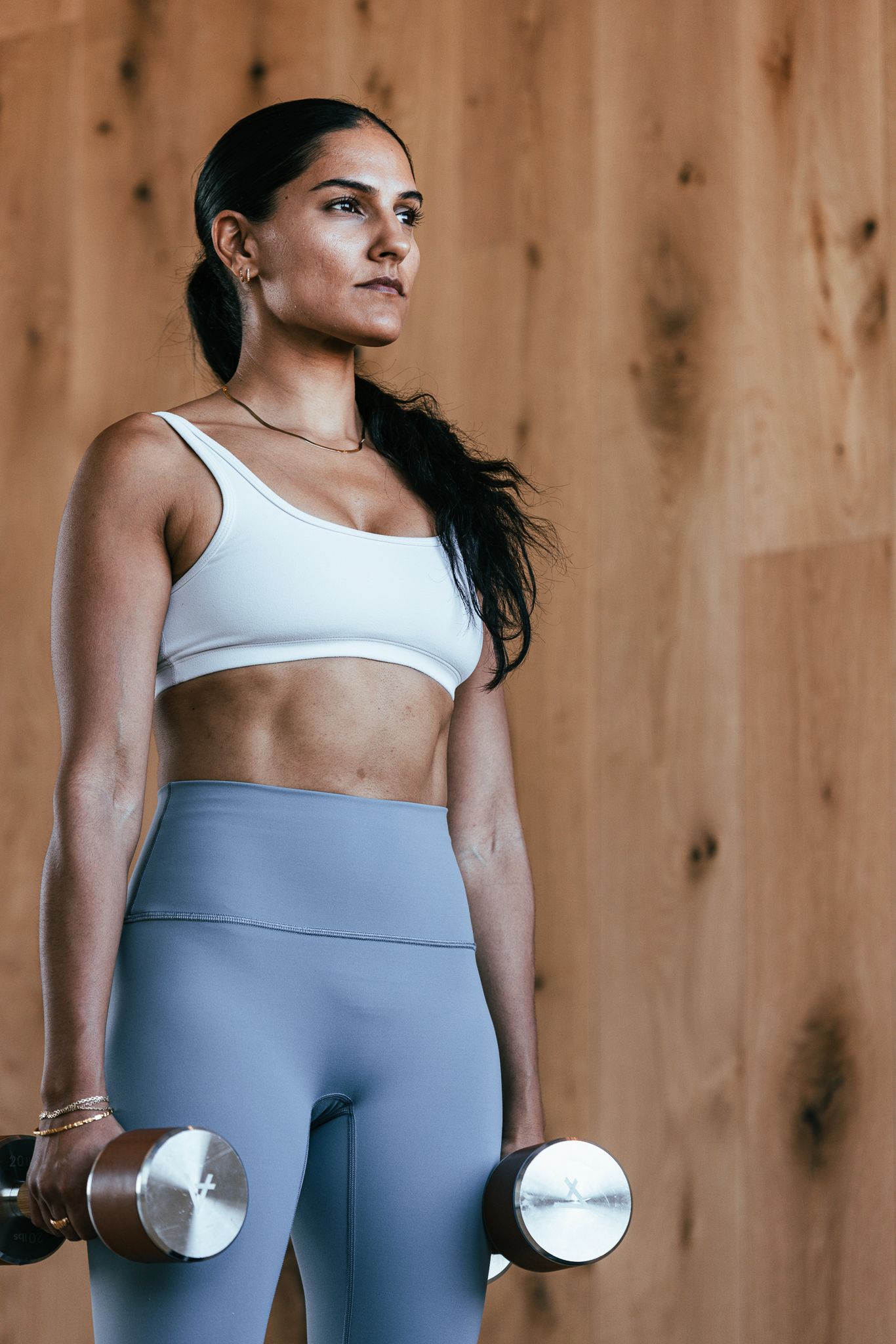 Fit woman in white sports bra and gray leggings holding dumbbells during strength training workout.