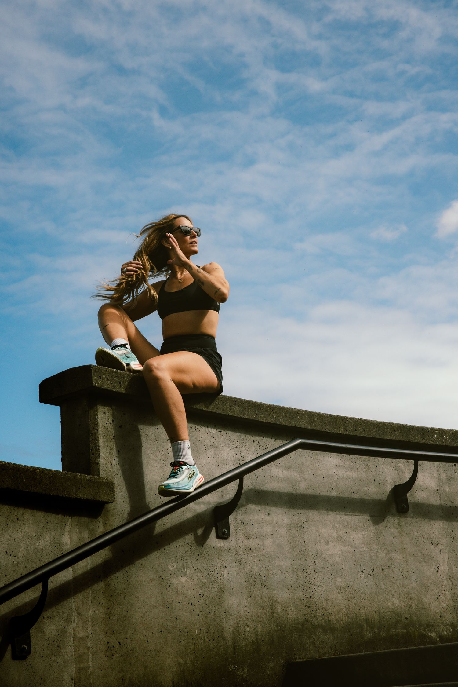 Female athlete in black sports bra and shorts sitting on a concrete wall against a cloudy blue sky.