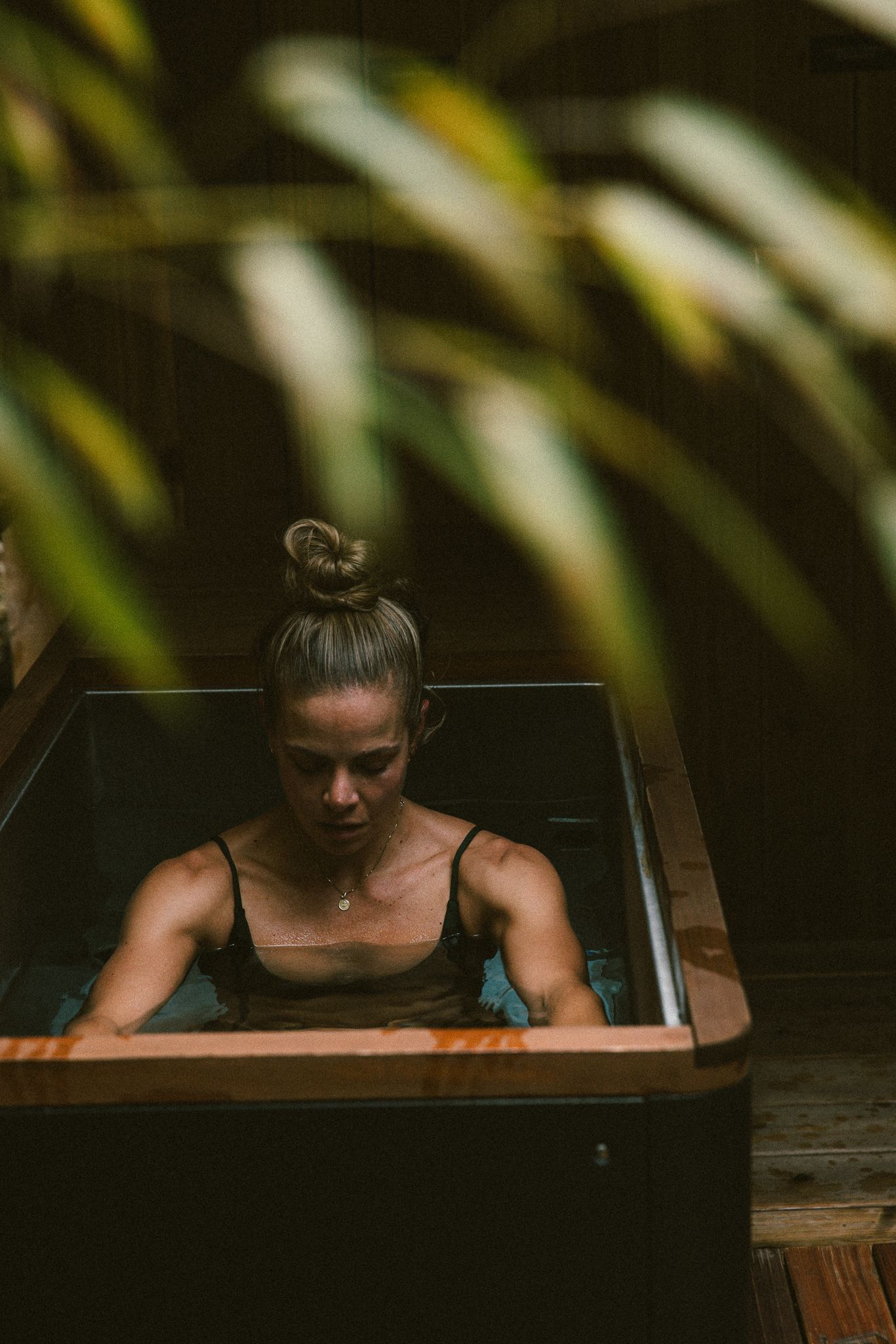Woman relaxing in a wooden cold plunge surrounded by tropical plants in a serene outdoor spa setting.