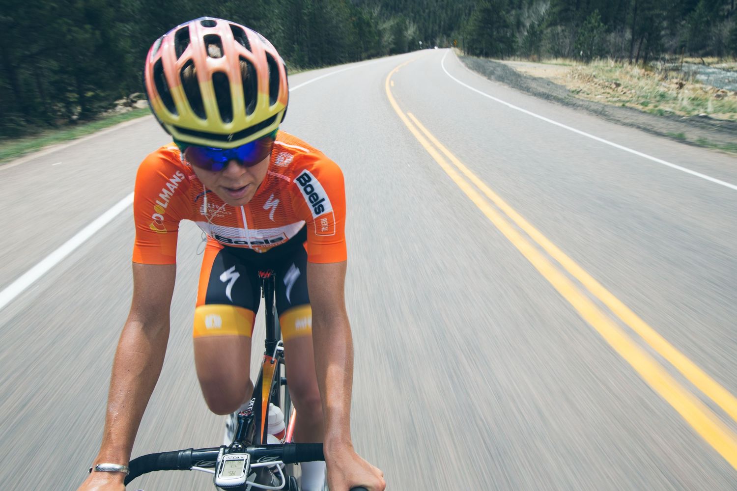 Cyclist in orange jersey and yellow helmet rides aggressively on open road during a competitive cycling race.