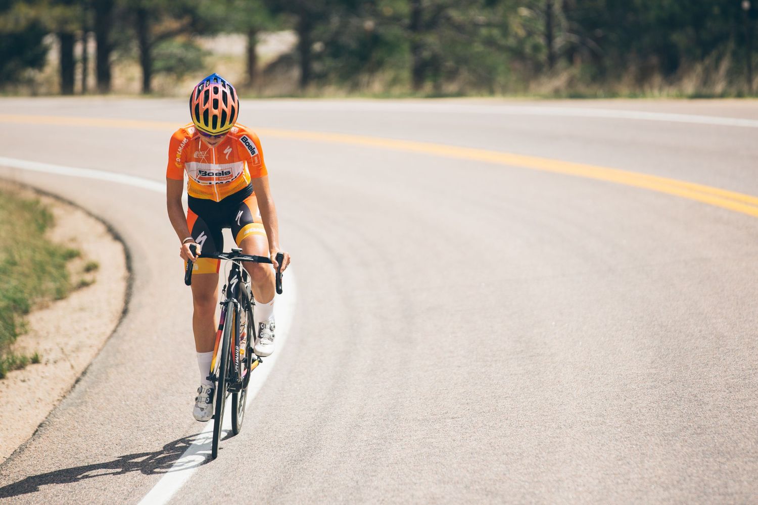 Cyclist in orange jersey and helmet riding a road bike on an open highway surrounded by trees.