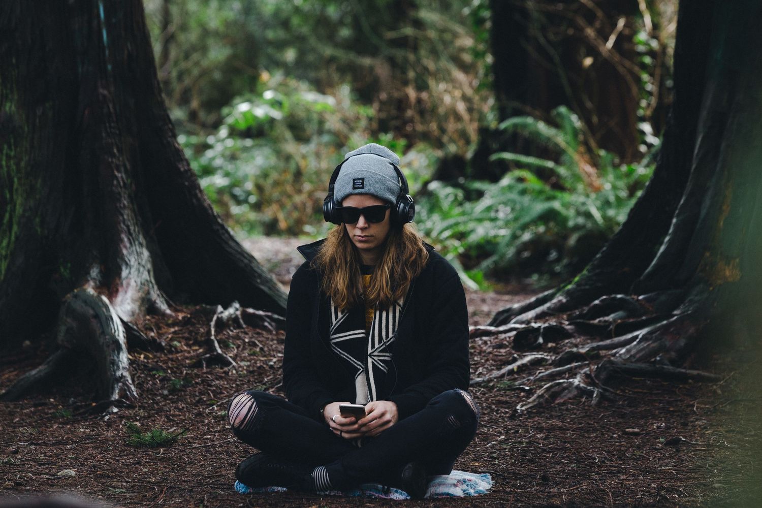 Woman in gray beanie and headphones sitting cross-legged in a lush forest surrounded by tall trees.