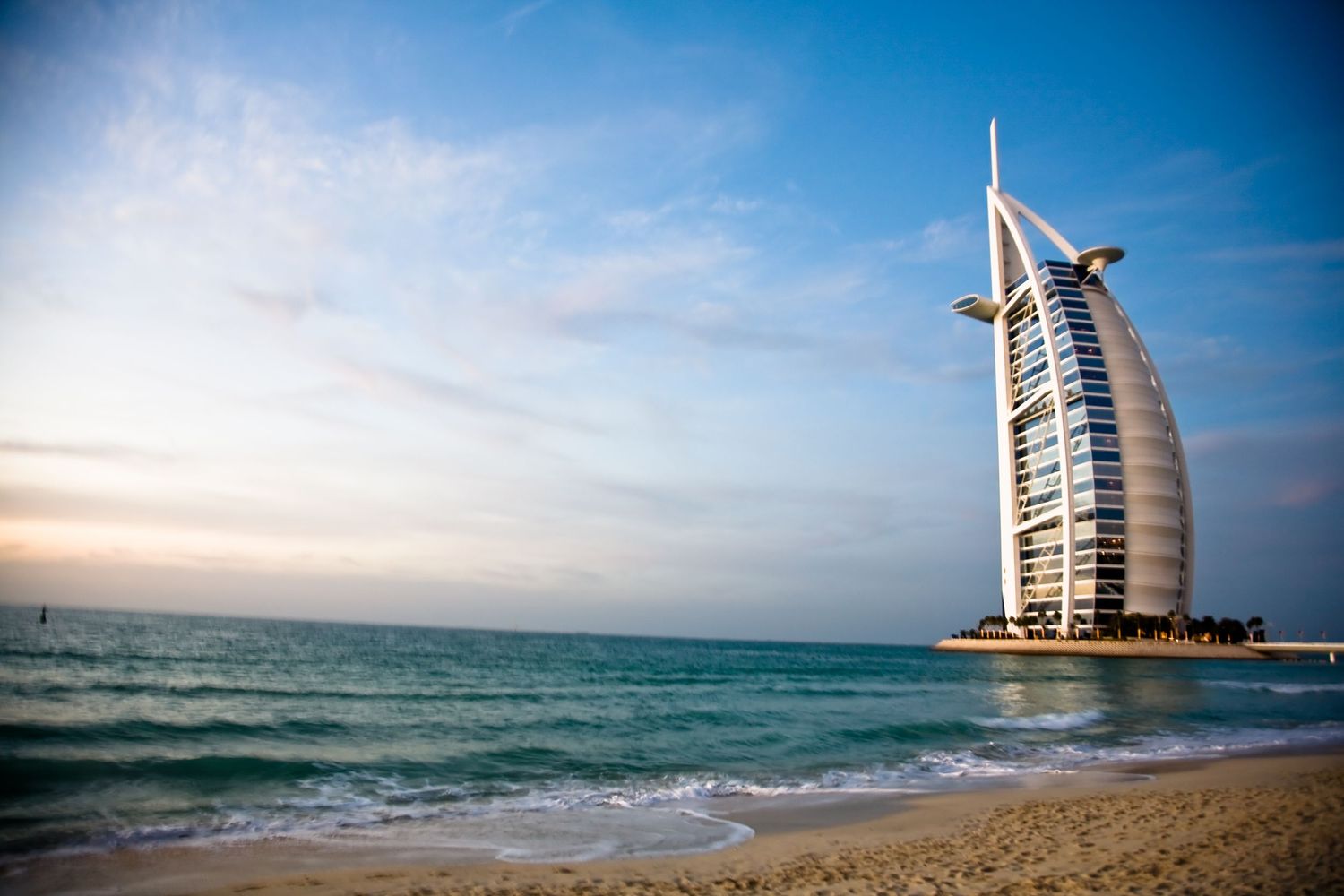 Burj Al Arab luxury hotel standing on artificial island with sandy beach and blue ocean waves in Dubai, UAE.