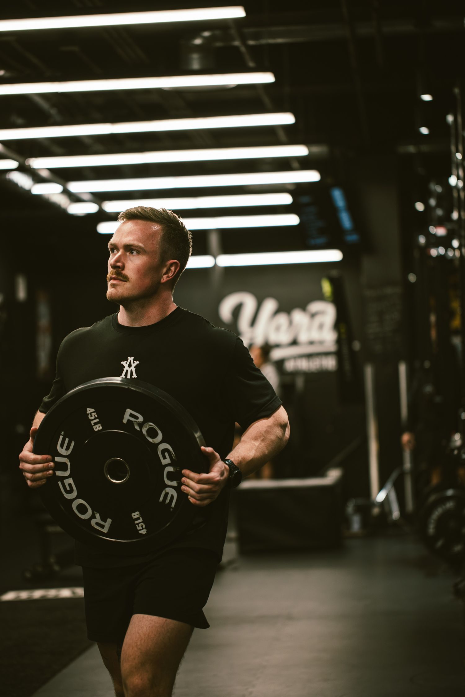 Fit man carrying a Rogue weight plate in a dark gym with neon lights.