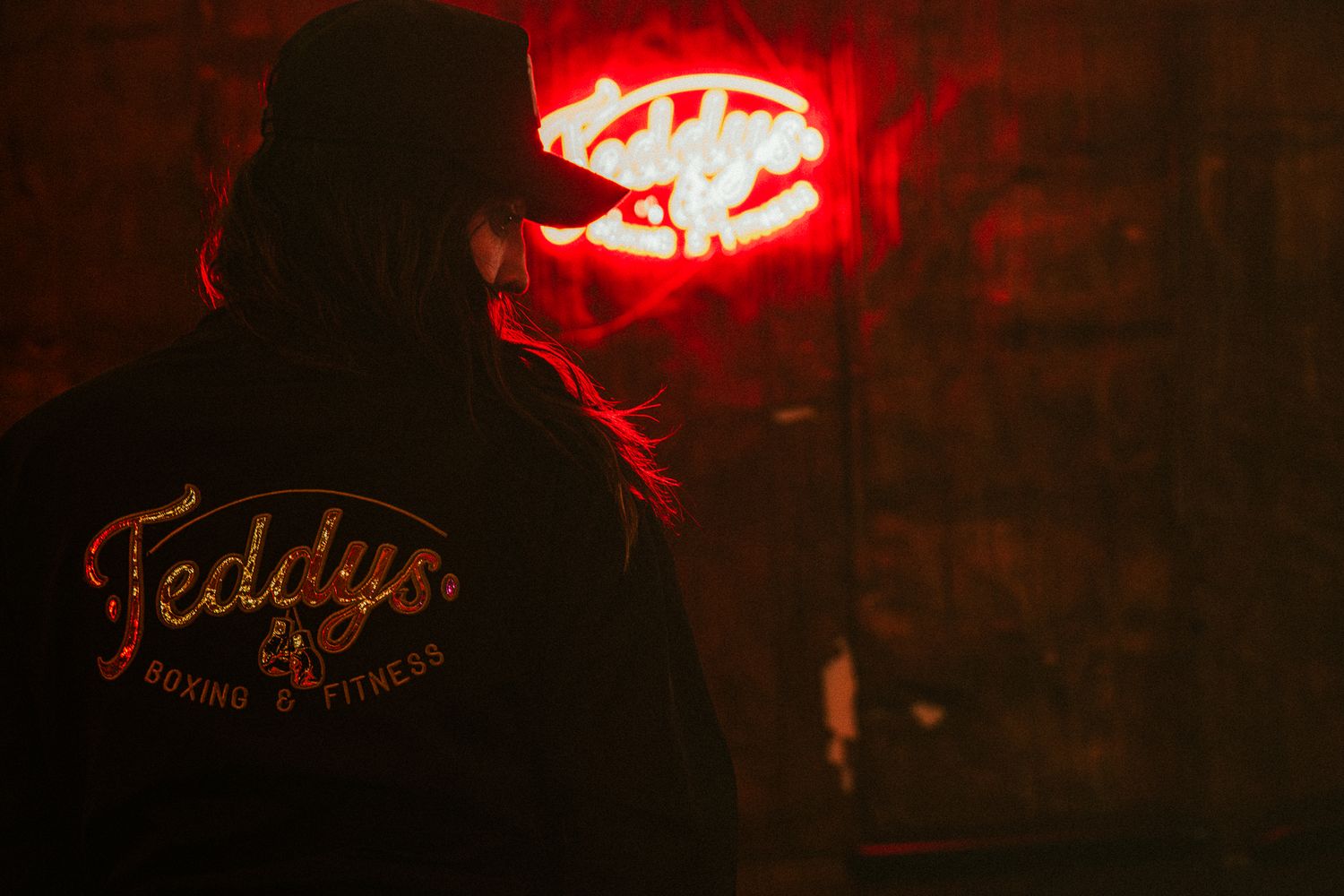 Person wearing Teddy's Boxing and Fitness jacket standing before a glowing red neon sign in dark gym.