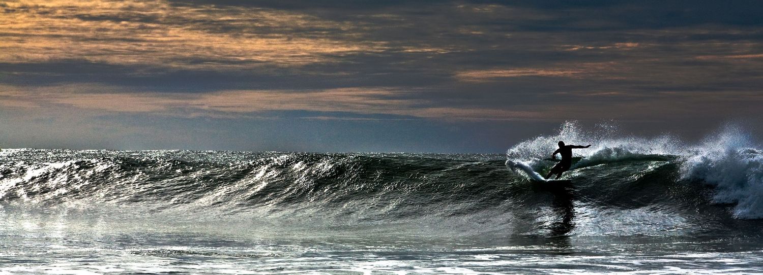 Silhouette of a surfer riding a large wave at sunset with dramatic cloudy sky in the background.