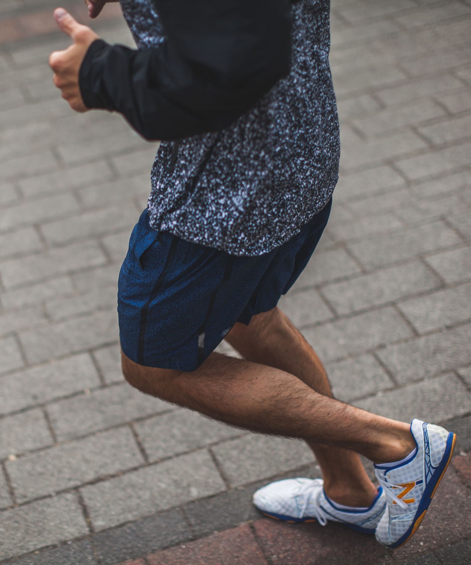 Runner in navy shorts and blue New Balance sneakers mid-stride on a brick path wearing a speckled dark hoodie.