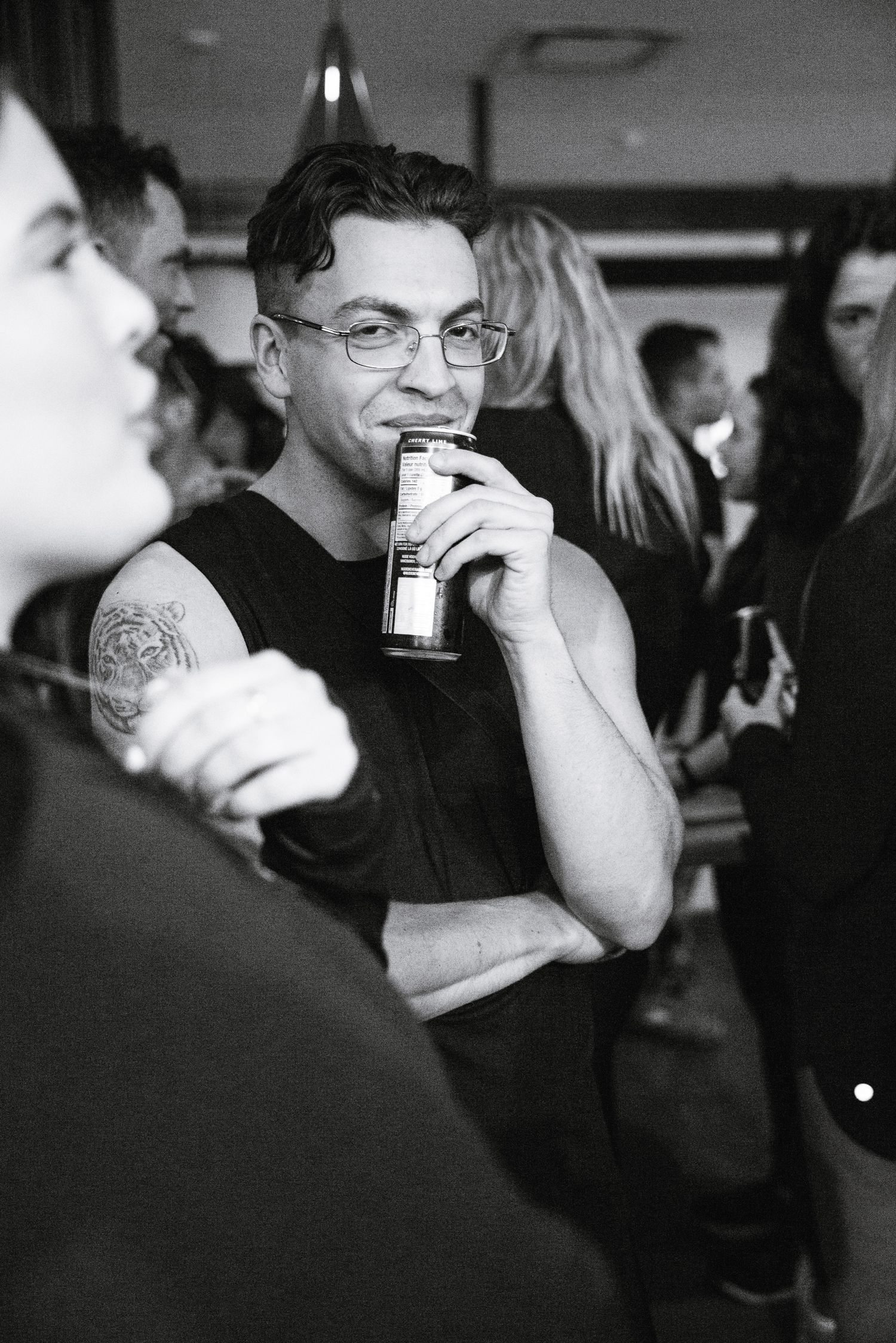 Black and white photo of tattooed man drinking from a bottle at a crowded indoor social event.