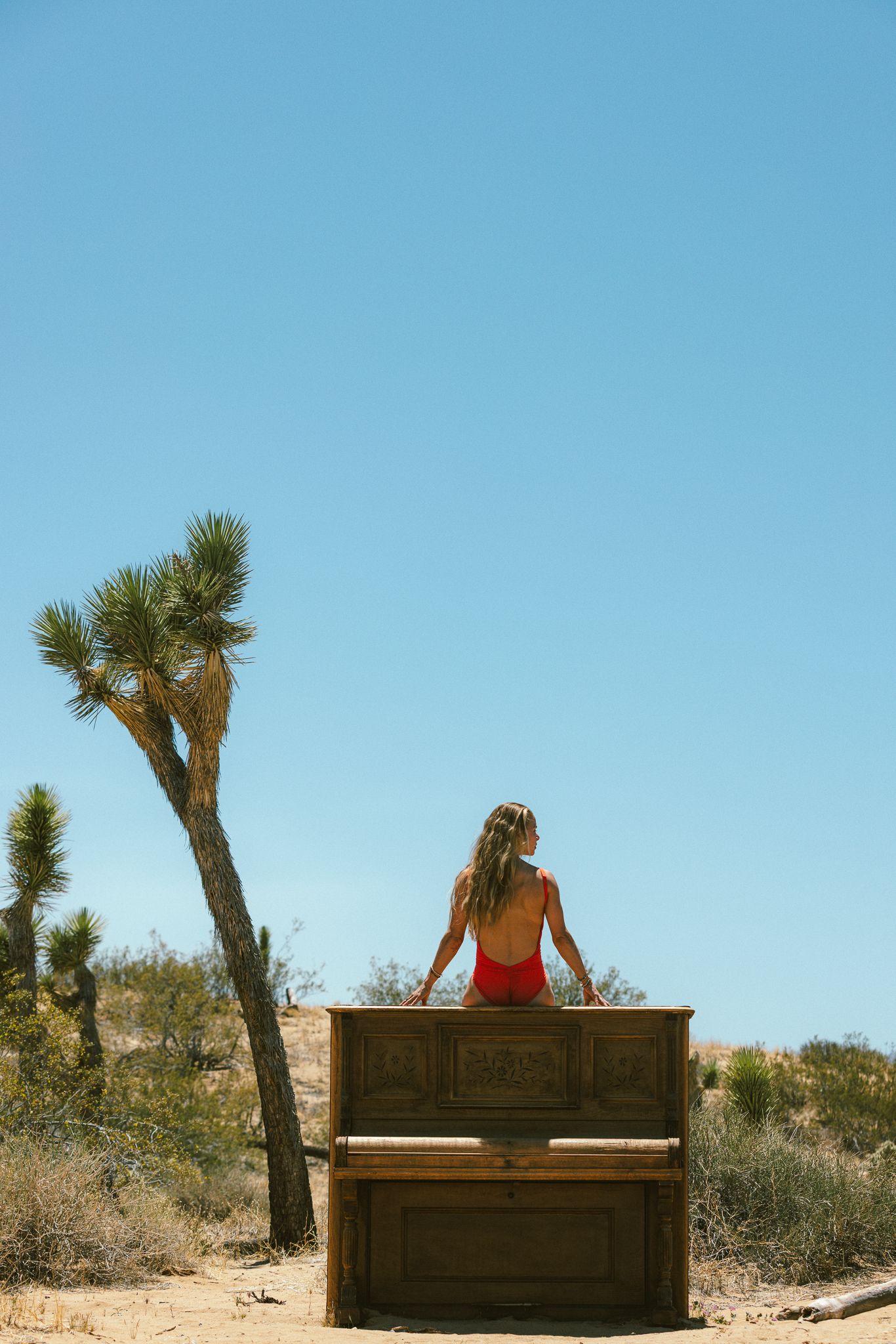 Woman in red swimsuit sitting on a vintage piano in desert landscape.