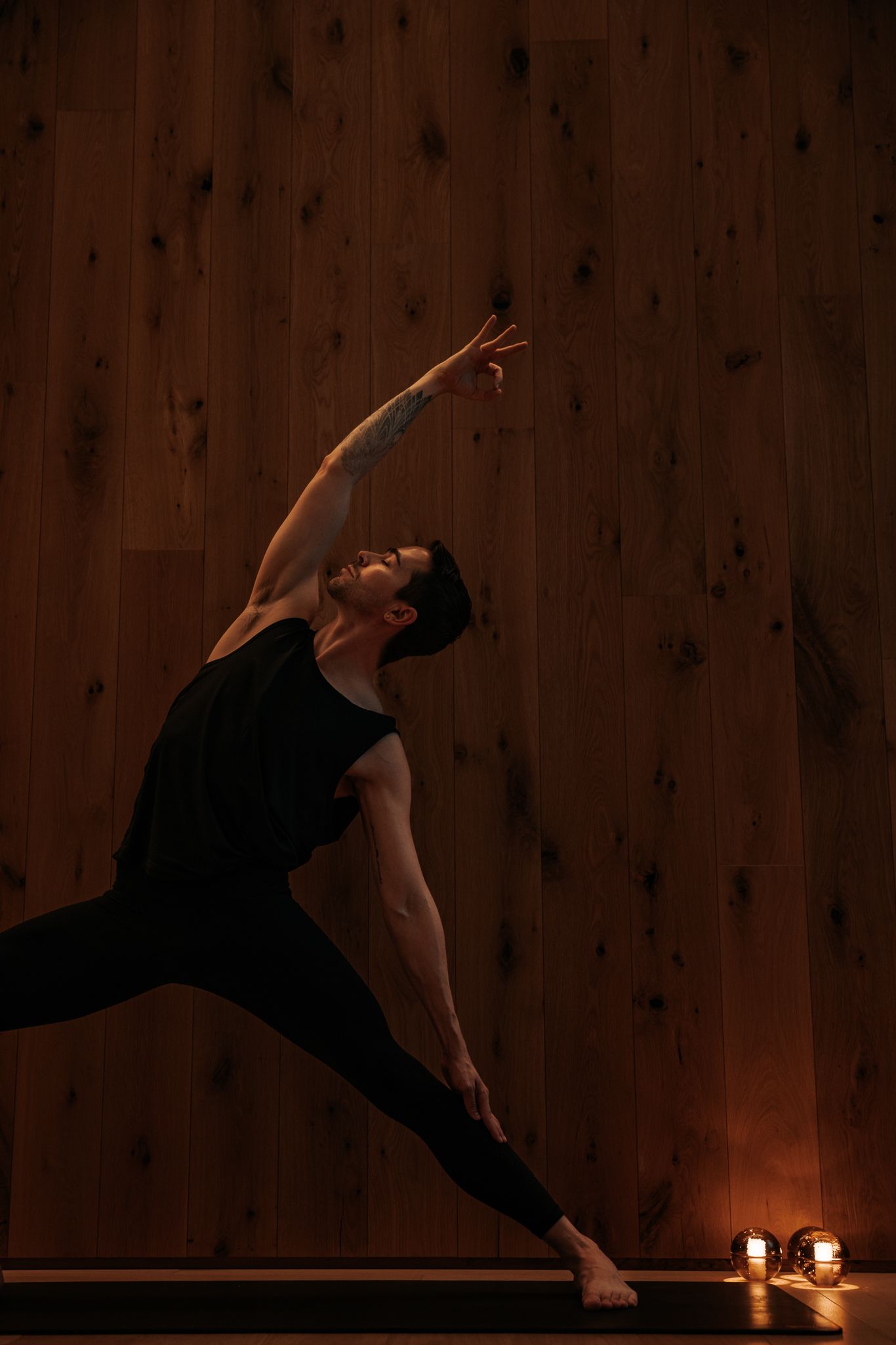 yogi in black performs a wide-legged side stretch pose by candlelight on a wooden floor.