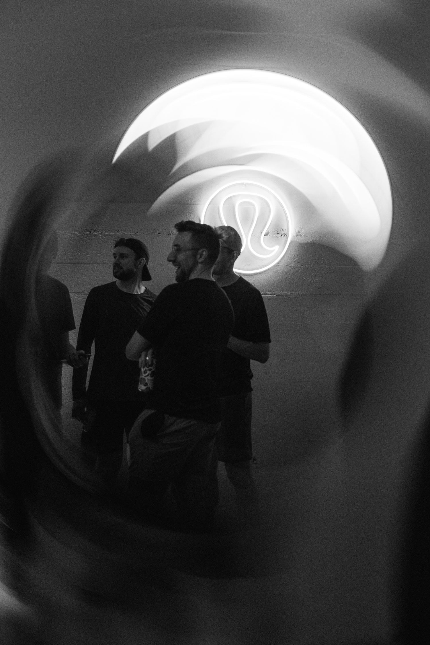 Black and white photo of two men in black shirts viewed through a circular tunnel with a neon logo glowing in background.
