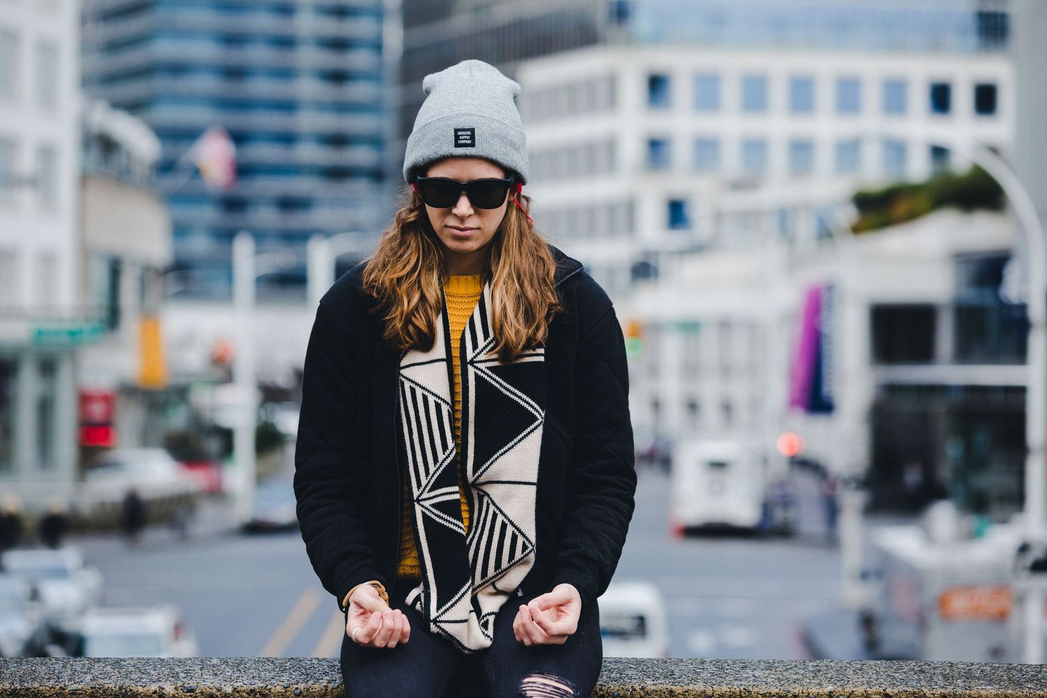 Young woman in gray beanie, sunglasses, and black jacket with geometric print sits on urban ledge.