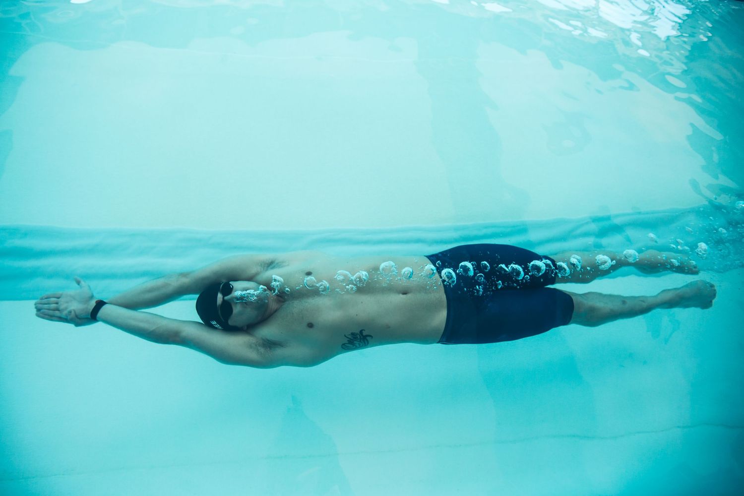 Male swimmer gliding underwater in a pool, wearing goggles and dark swim trunks, captured from below in clear blue water.