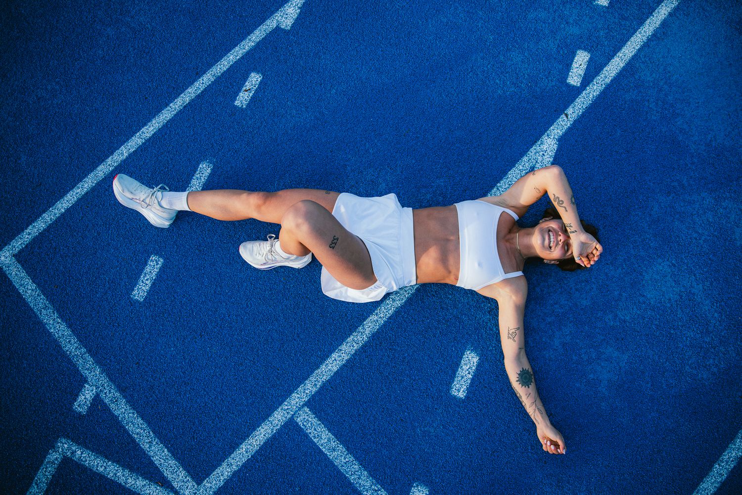 Overhead view of athletic woman lying on blue track in white workout outfit with tattoos.