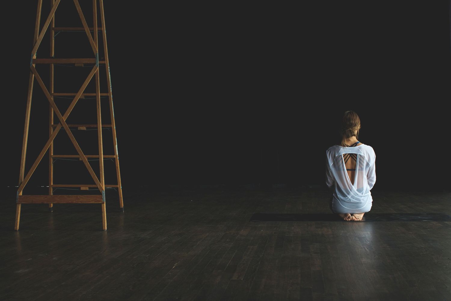 Woman sitting on floor in white open-back top beside wooden ladder in dark studio.